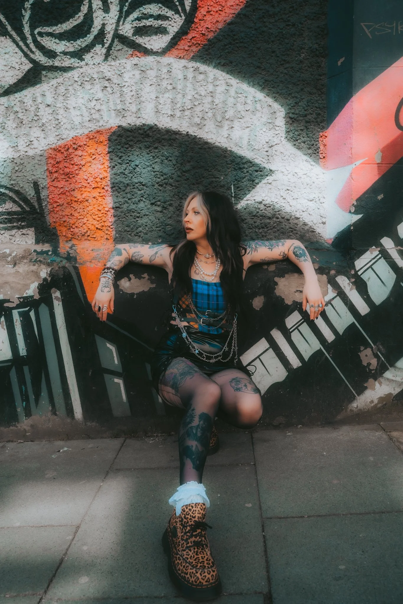 A woman with long dark hair and tattoos sits against a graffiti-covered wall, wearing a blue plaid top, layered necklaces, and leopard-print boots. Alternative styles, Alt Girl, Goth girl, punk