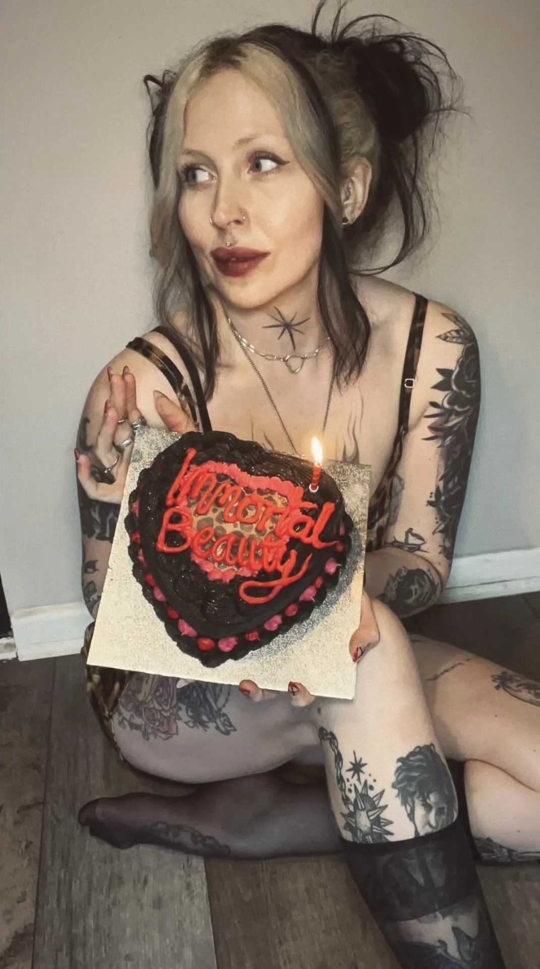 A woman with multiple tattoos and dyed hair holding a heart-shaped cake with a single lit candle and the words 'Immortal Beauty' written in red icing. Leopard print cake, Girl with tattoos, Goth girl, Alternative style, Alt girl