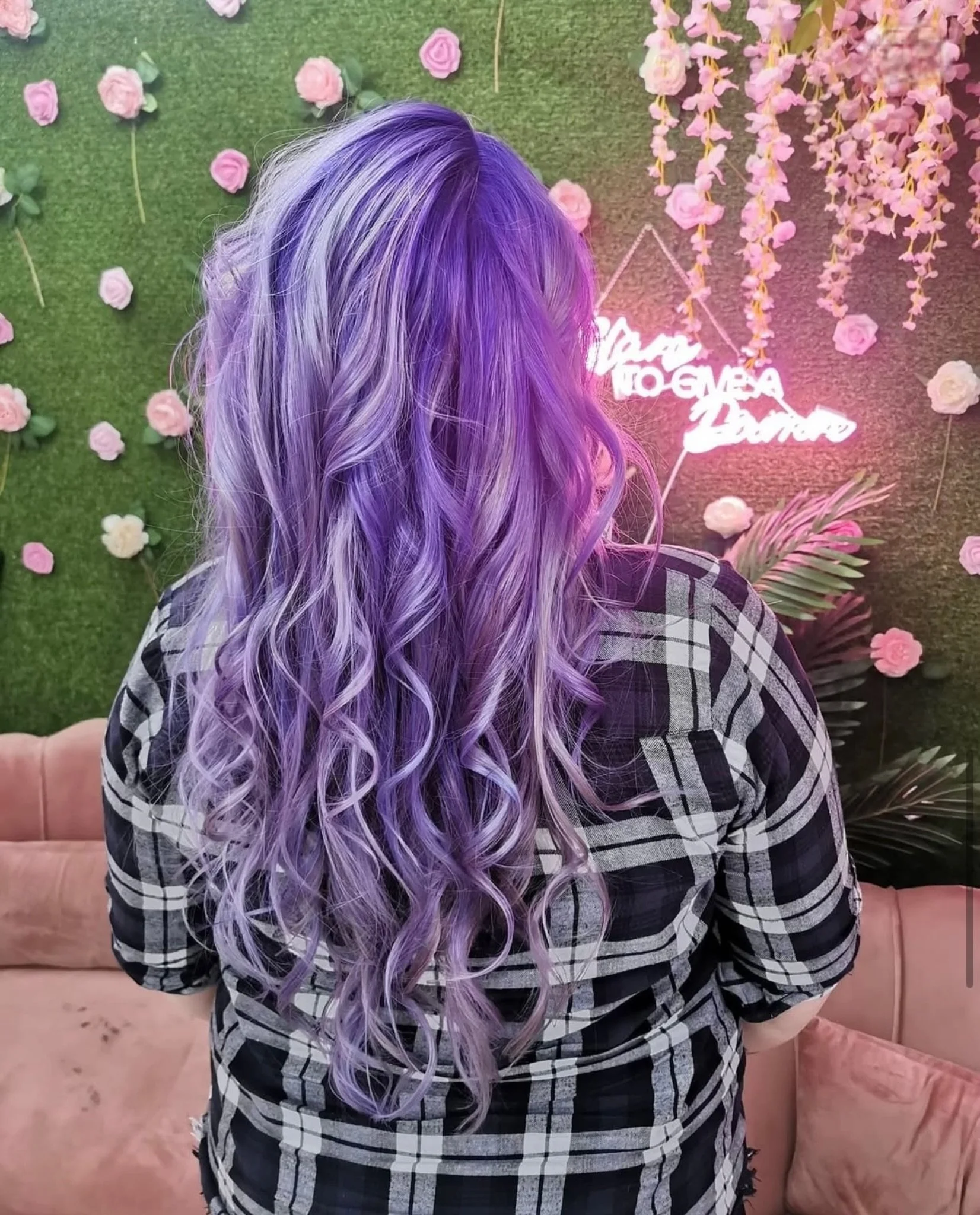 Back of a person with long, wavy hair dyed in shades of purple and lavender, wearing a black and white plaid shirt, standing in front of a green wall decorated with pink and white artificial roses and flowers, with a neon sign and a pink cushioned bench.