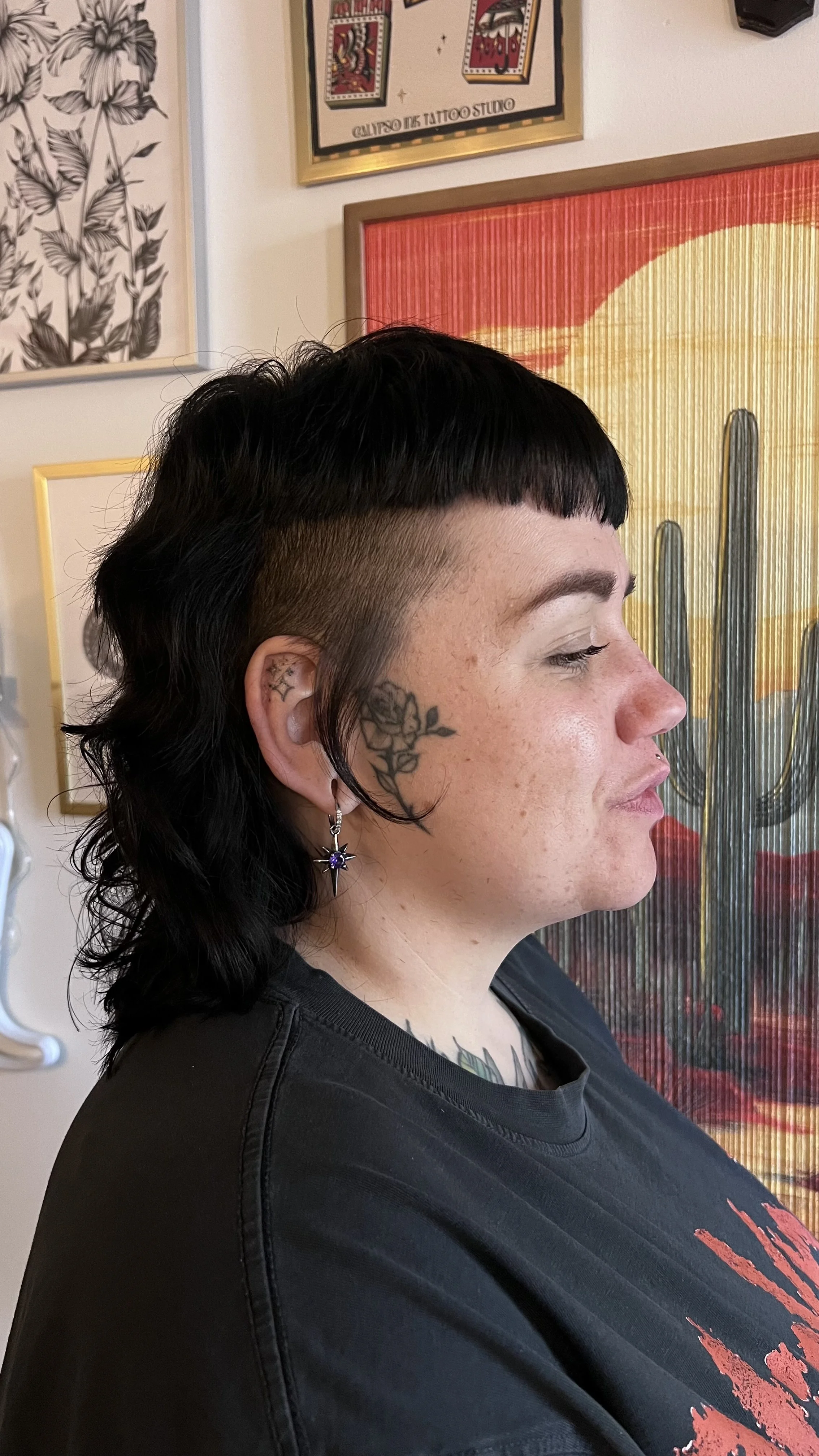 Profile of a woman with short black hairstyle with a shaved side, tattoos on her face and neck, and wearing earrings, in front of wall art including a cactus print and tarot card images. Mullet, Alt style, punk, goth girl, alt girl, face tattoo