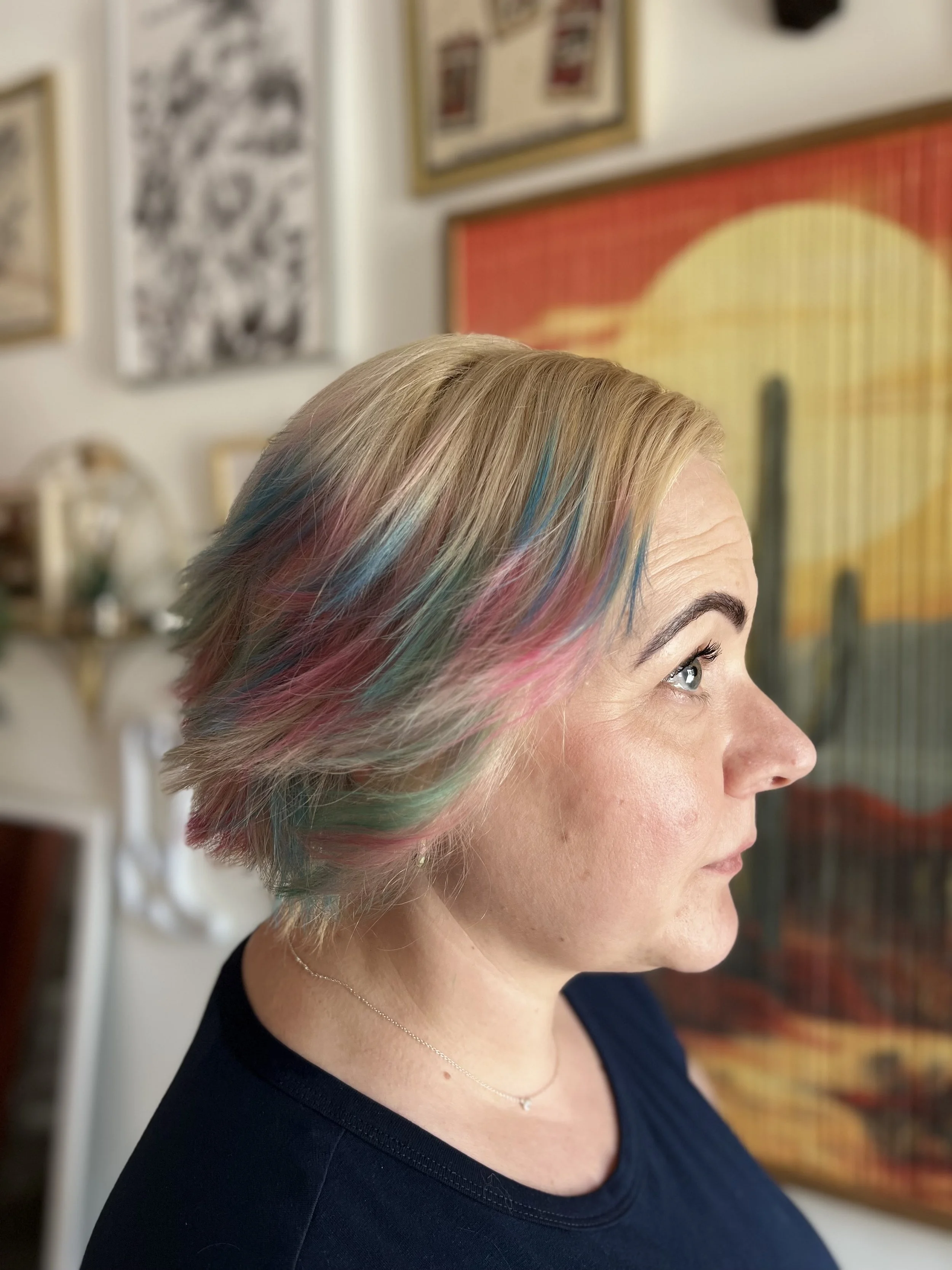 A woman with short blonde hair with streaks of blue and pink, wearing a navy blue top and a delicate silver necklace, standing in a room with art and pictures on the walls.