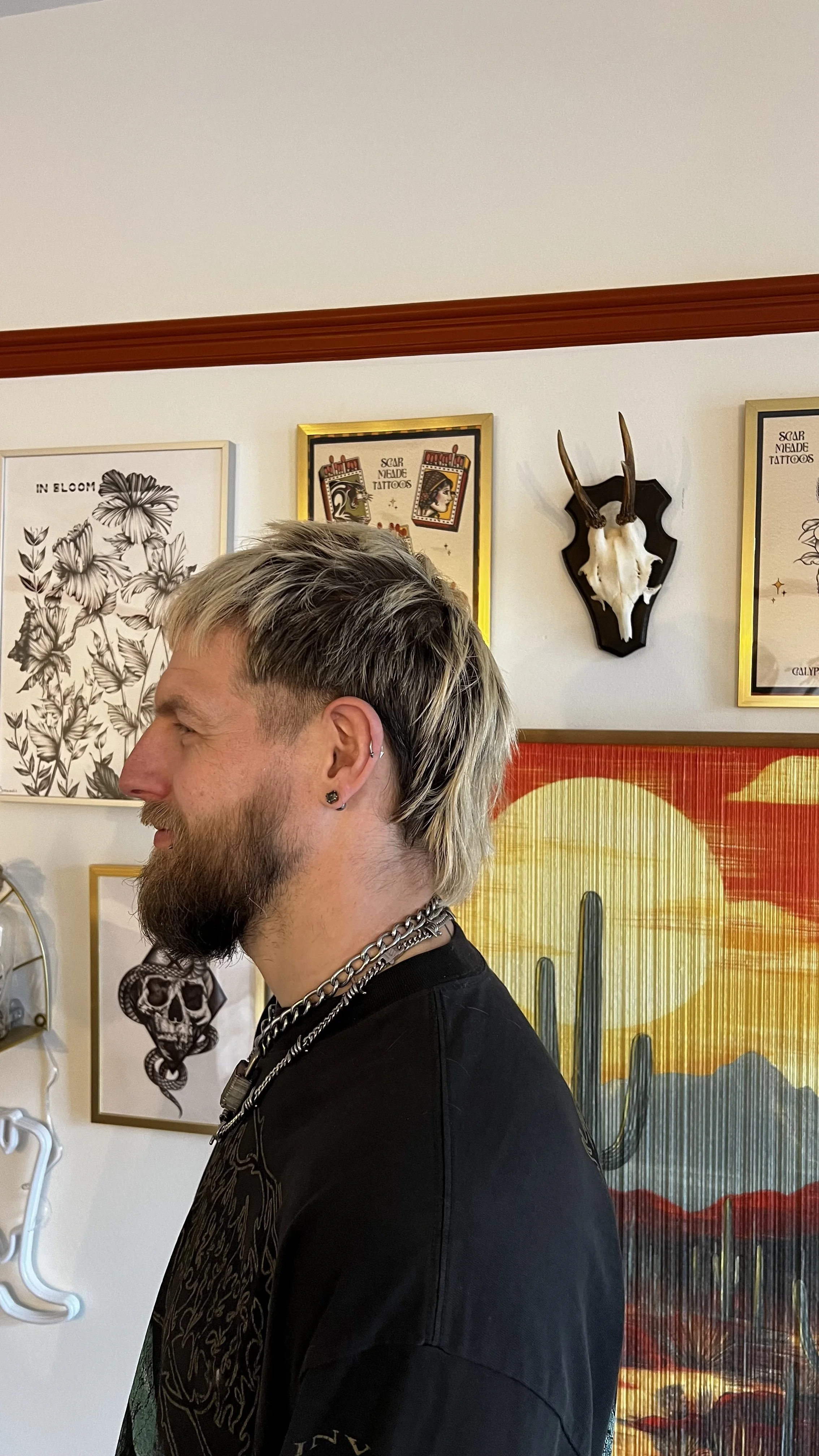 Profile of a man with blonde, black and white hair and a beard, wearing earrings and a chain necklace, standing indoors with artwork and mounted animal skulls on the wall behind him. Mullet, alt style, punk, hair stylist, newark, nottingham