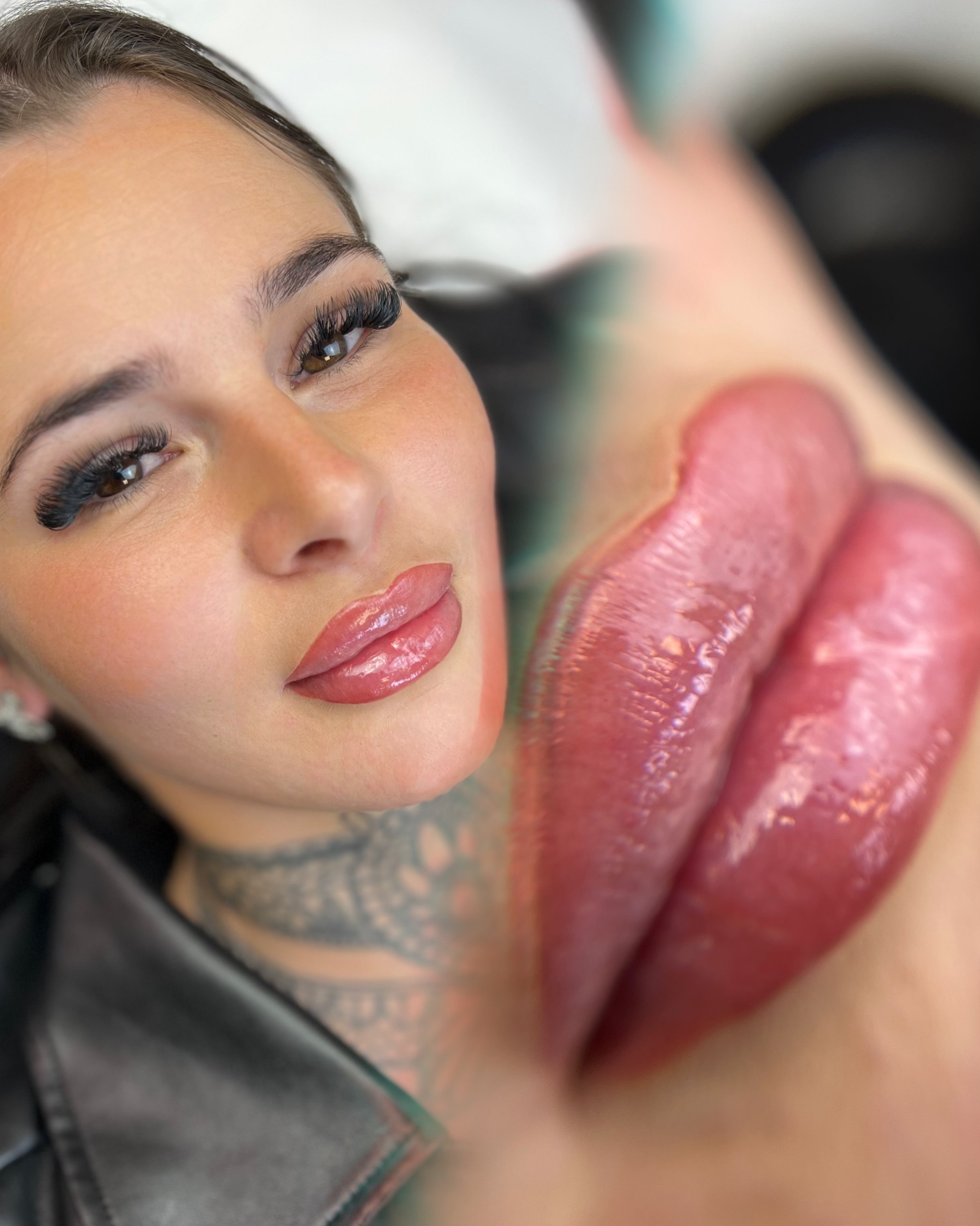 Close-up of a woman's face with makeup, focusing on her eyes and lips, with a blurred figure in the background. Lip Blush, Girl with tattoos, alt girl, goth girl, alternative style
