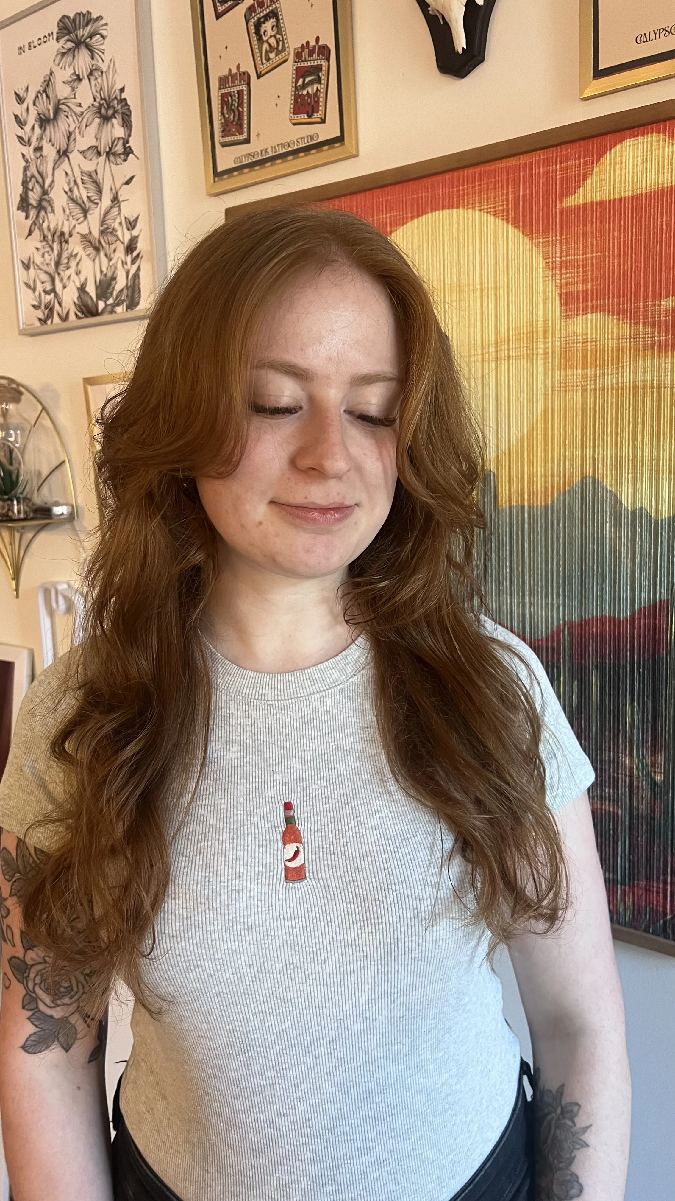 A woman with long, wavy red hair, closed eyes, and a content smile, wearing a light gray t-shirt with a small graphic of a red lipstick and a lip gloss tube. She has tattoos on her left arm and is standing indoors against a colorful, patterned background with various framed art pieces on the wall.