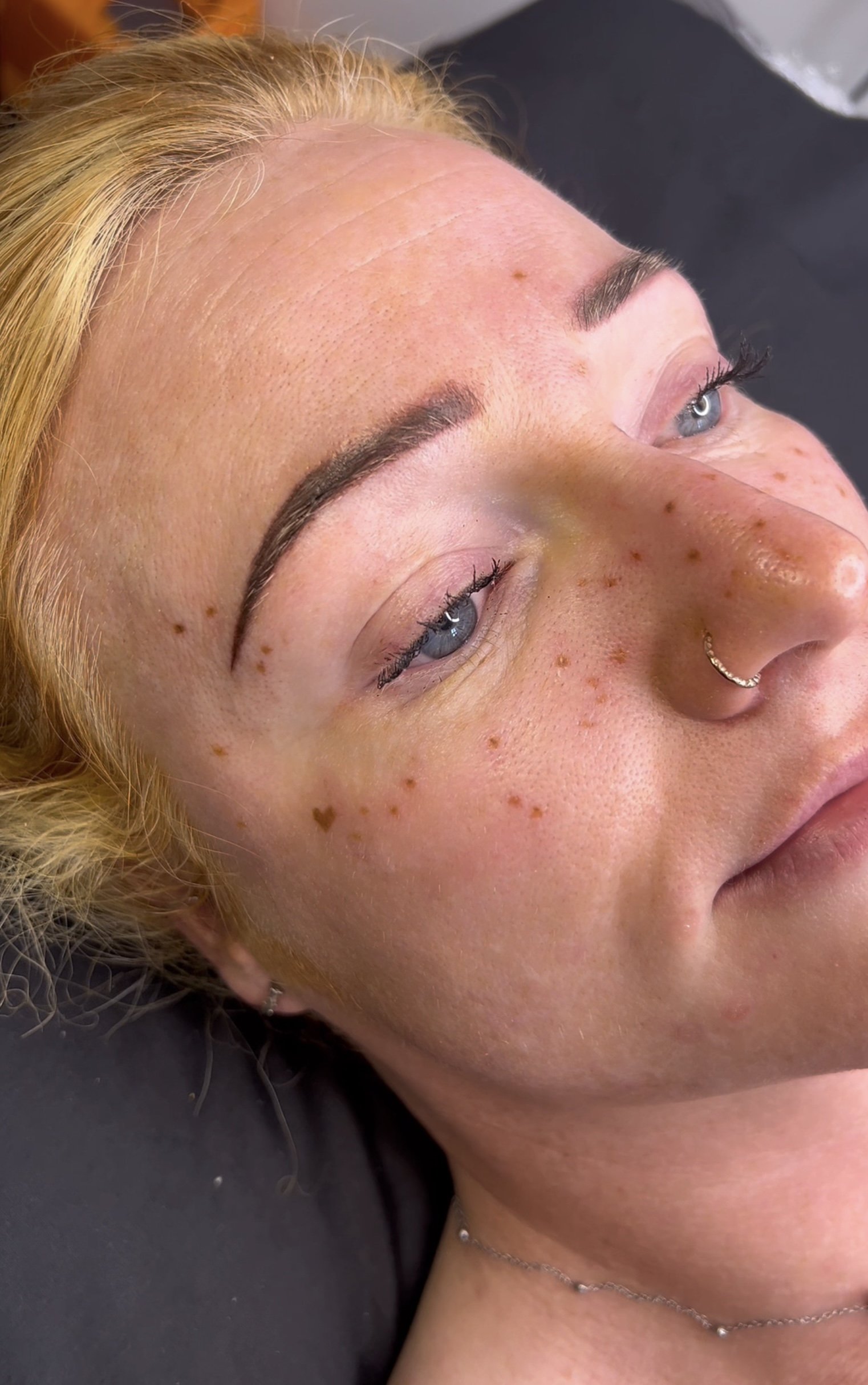 Close-up of a woman’s face with blonde hair, blue eyes, freckles, and a nose ring, lying on a dark surface. Tattooed freckles, heart freckle, freckle heart, permanent make up, pmu artist, nottingham, newark