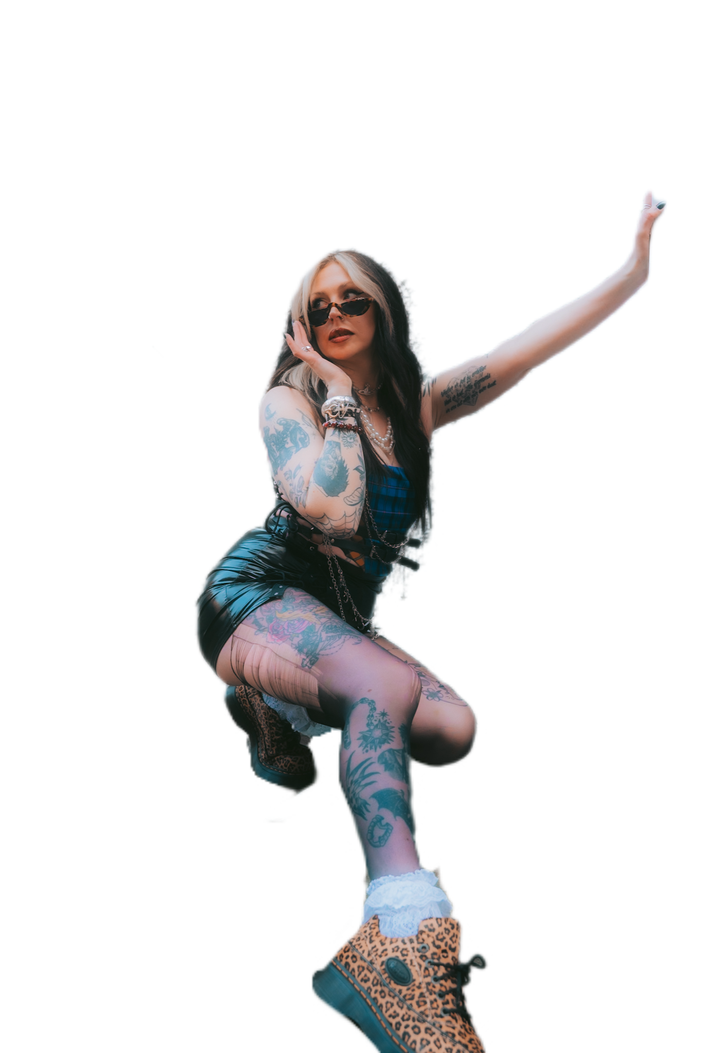 Woman with tattoos on arms and legs, black and platinum blonde hair, sunglasses, wearing a blue checkered top, black leather skirt, and leopard print boots, posing with one arm raised and the other hand near her face.