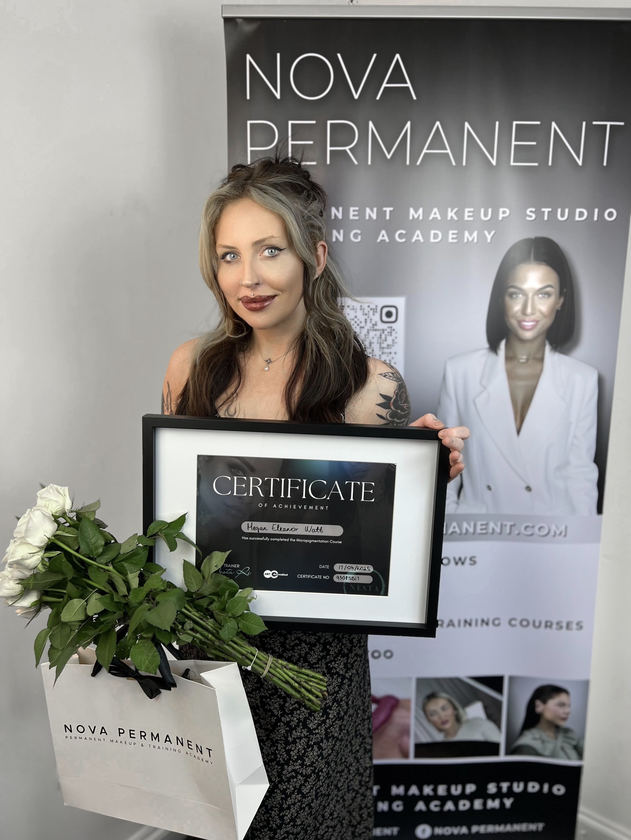 A woman holding a certificate of achievement from Nova Permanent Makeup & Training Academy, with a bouquet of white roses in a white bag from the same academy, standing in front of a promotional banner for the academy.