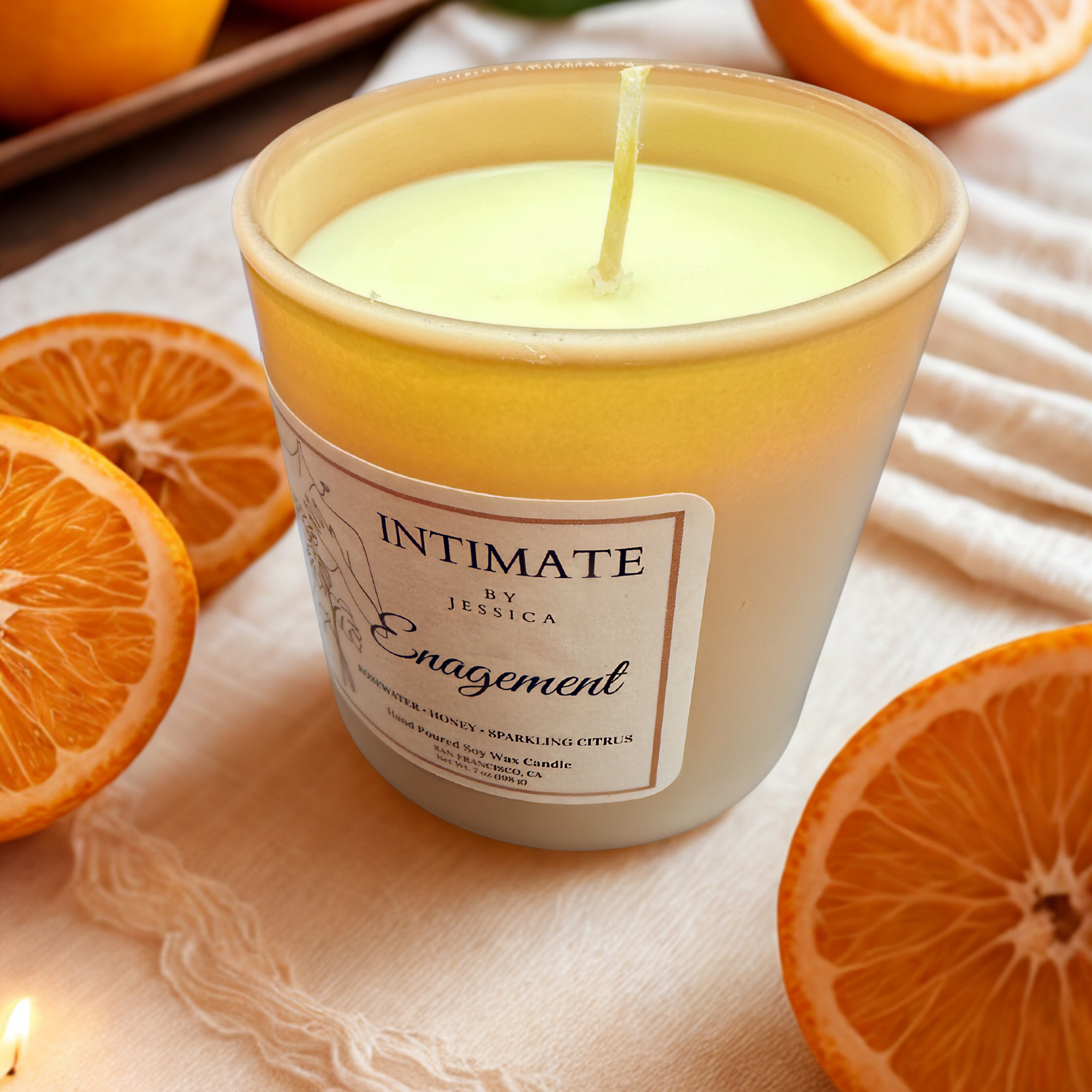Close-up of a lit candle in a yellow container with a label, surrounded by sliced oranges on a soft fabric surface.