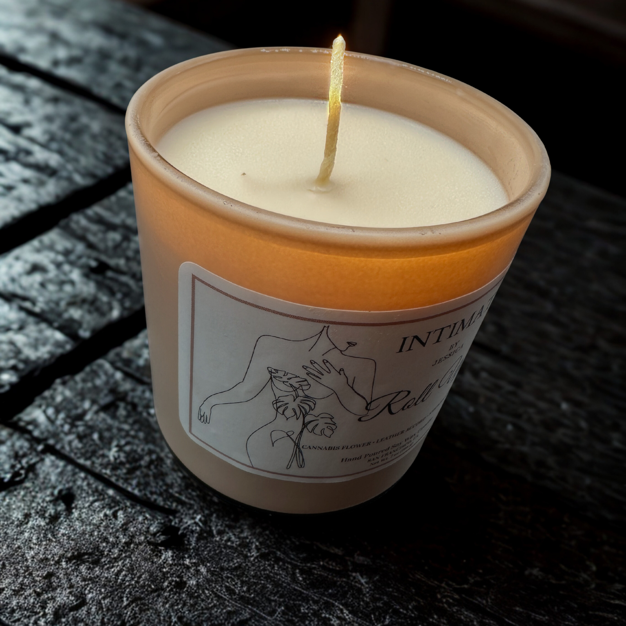 A lit candle in a beige container with a label that features an illustration of a cannabis plant. The candle is placed on a dark, textured surface.