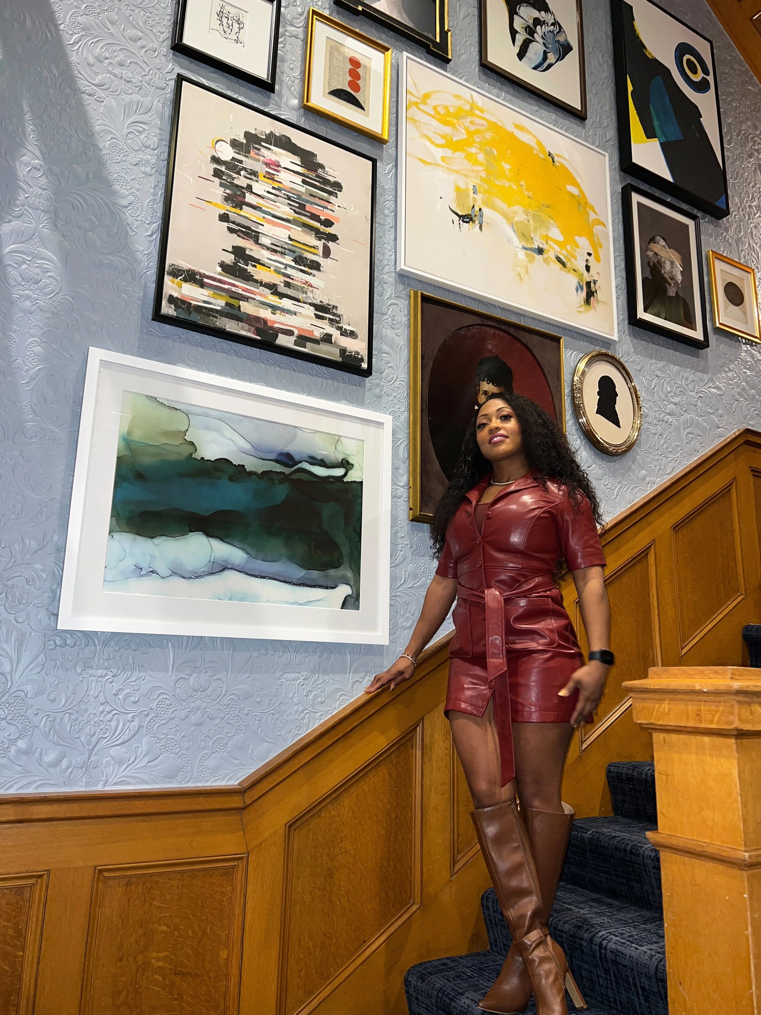A woman with curly dark hair wearing a red leather dress and tall brown boots standing in front of a gallery wall of various abstract and portrait artworks in a room with wooden paneling and textured gray wallpaper.