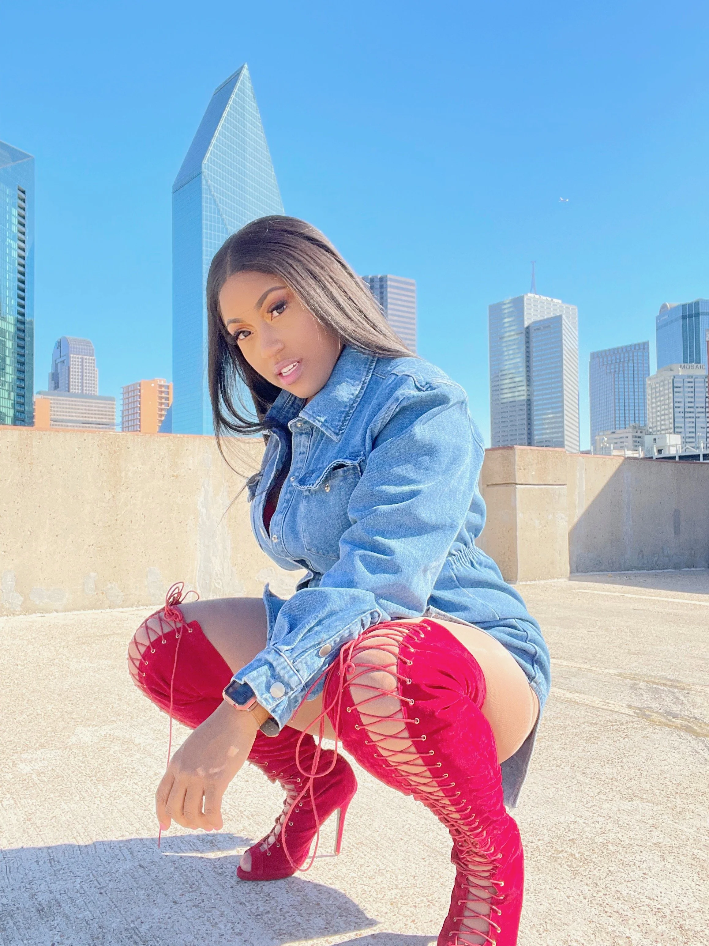 A woman is kneeling on a rooftop with the city skyline in the background, wearing red lace-up high-heeled boots, a denim jacket, and shorts.