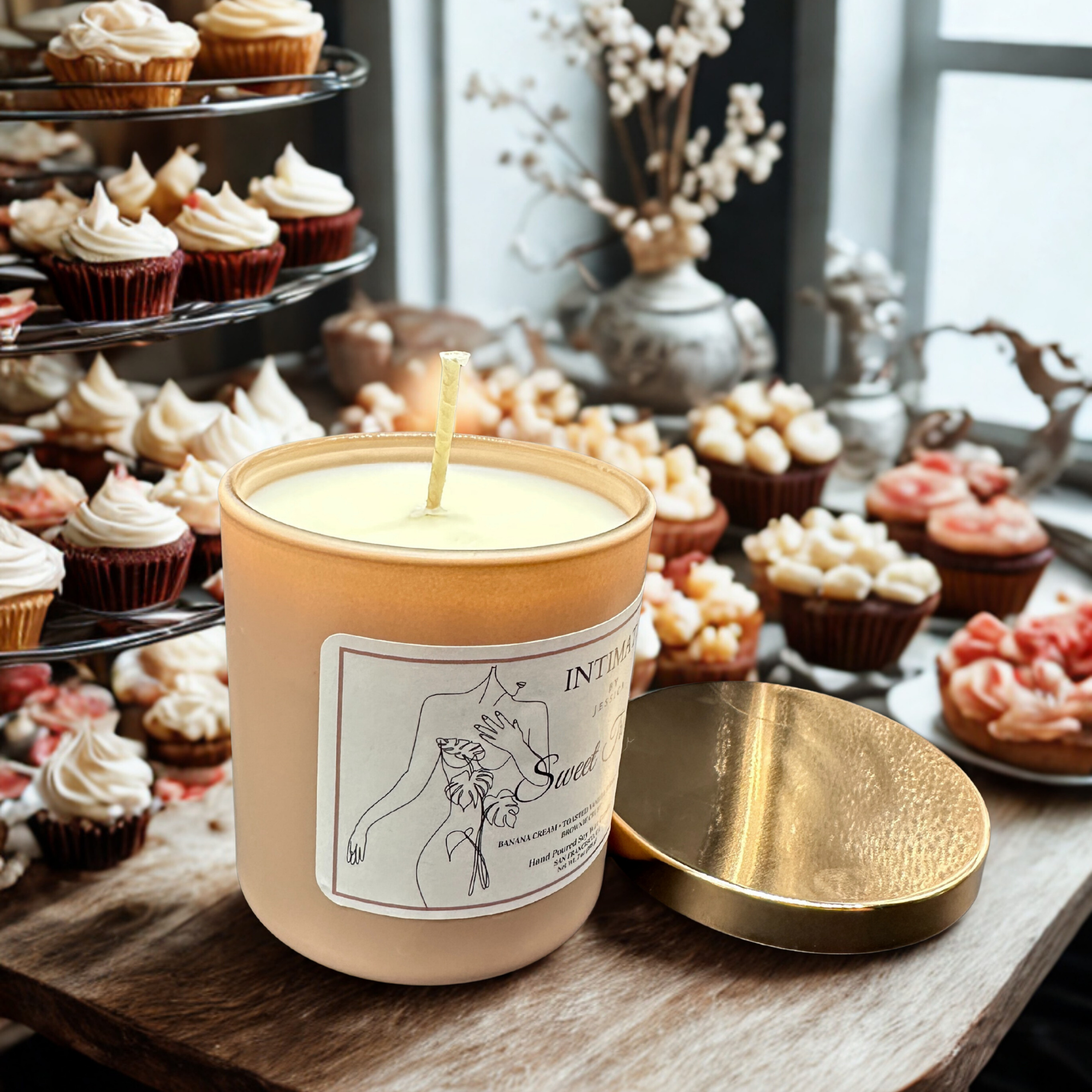 A beige cup with a label featuring a line drawing of hands holding a flower and text that reads 'Sweet'. The cup contains a beverage with a straw. In the background, there are cupcakes with white frosting on a metal tiered serving tray, and a rustic vase with white cotton stems, all placed on a wooden surface.