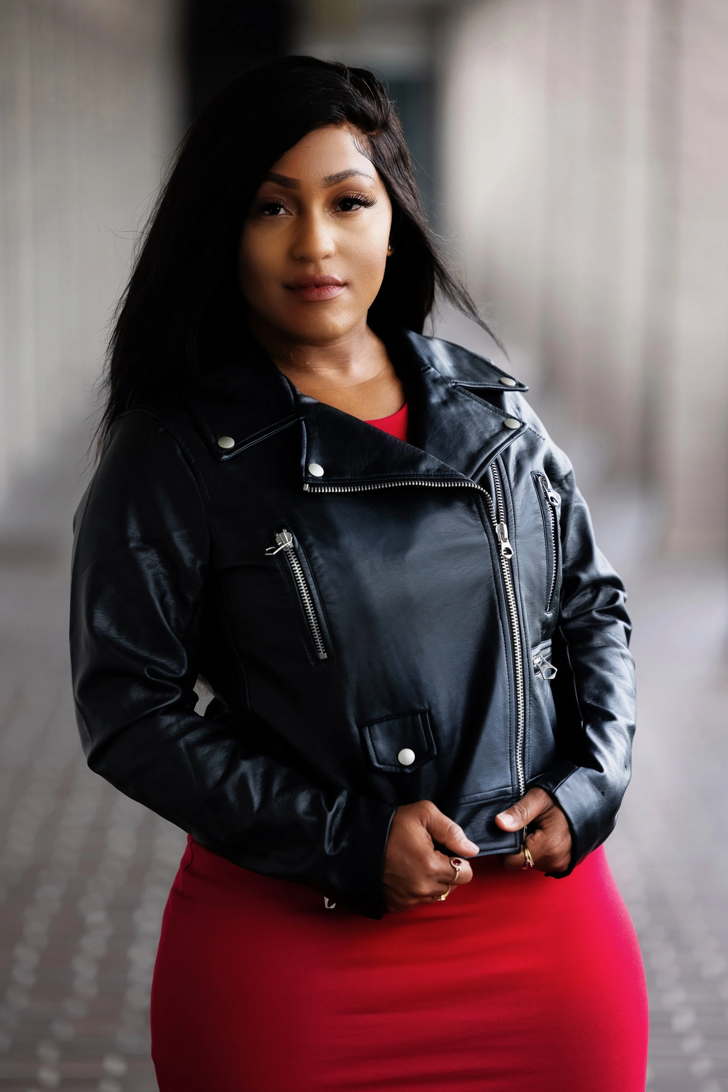 A woman with medium skin tone and long dark hair, wearing a black leather jacket and a red dress, standing outdoors on a paved walkway.