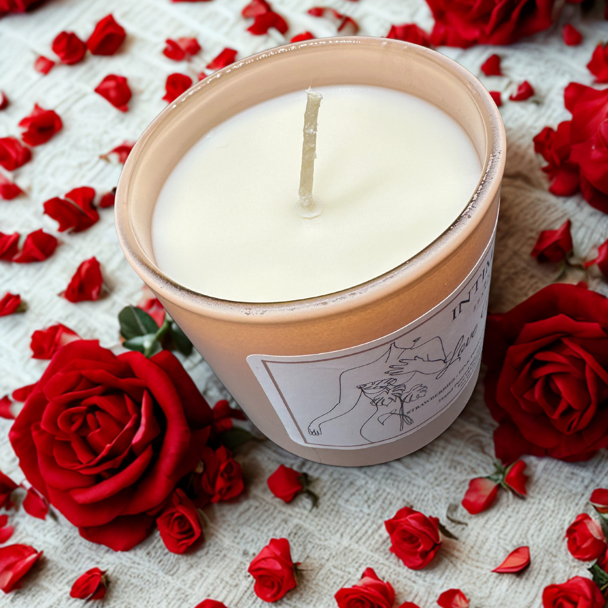 A white candle in a beige container surrounded by red rose petals on a textured beige surface.