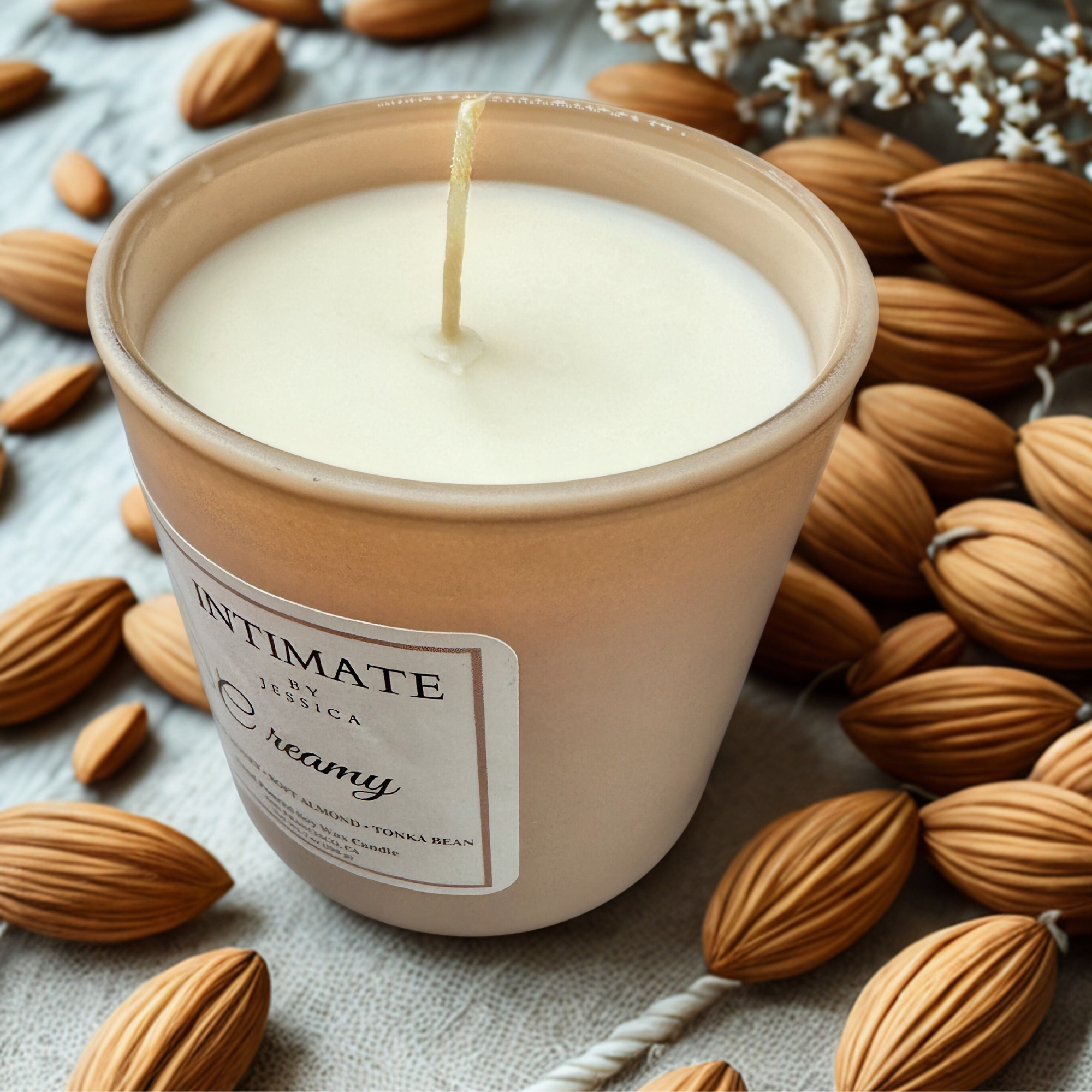 A candle in a beige container surrounded by almonds and white flowers on a textured surface.