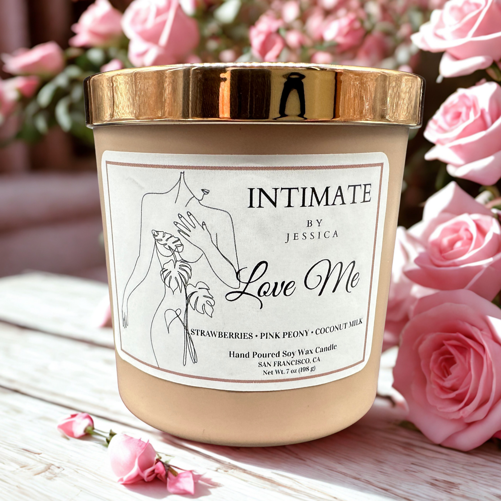 A beige candle with a gold lid, placed on a white surface, surrounded by pink rose petals and pink flowers, with a partly visible label that reads Love Me and mentions scents like strawberries, pink peony, coconut milk, and a soy wax candle.