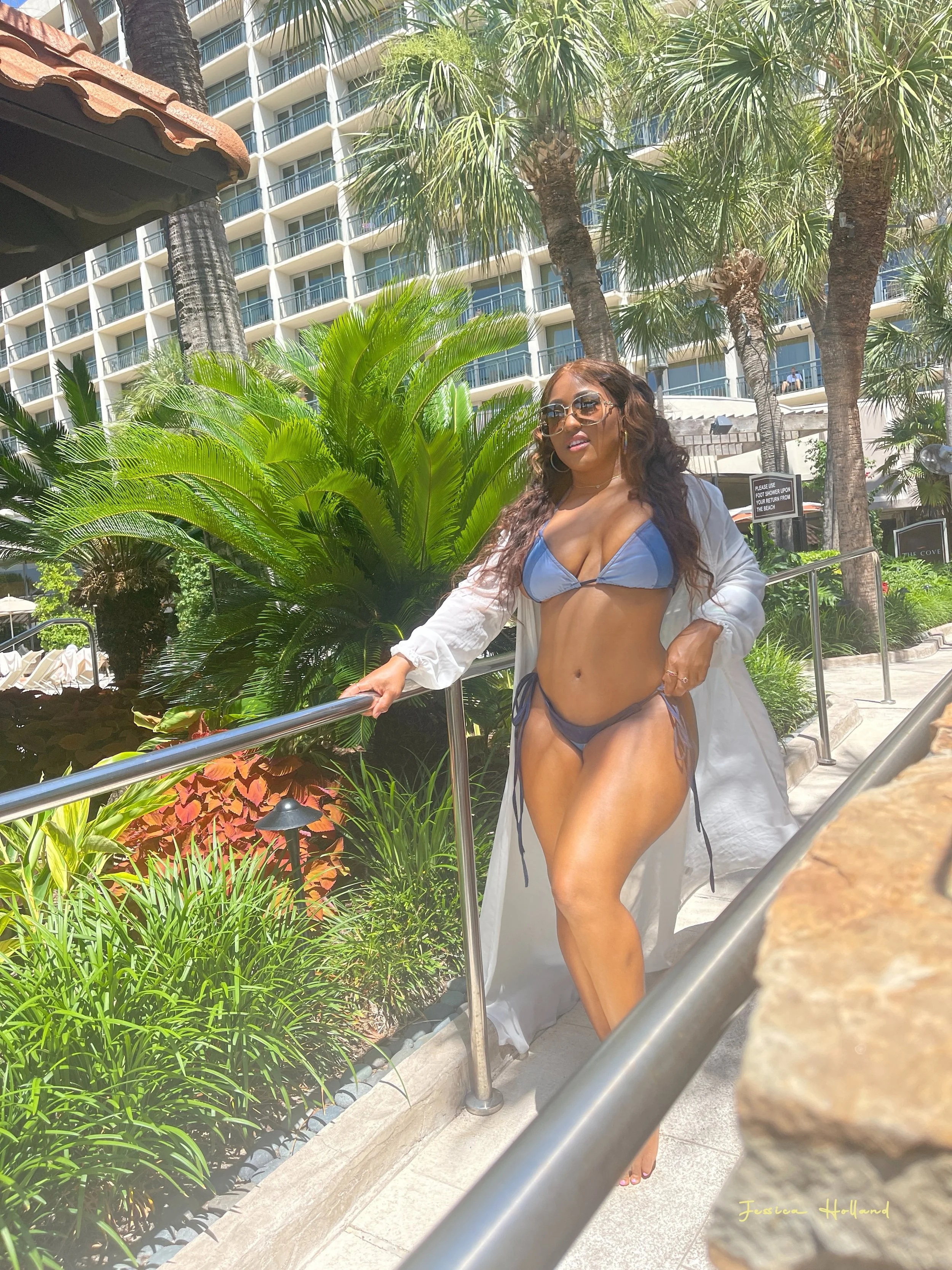 Woman in a blue bikini top and matching bottom, sunglasses, and a white cover-up, standing outdoors near tropical plants and palm trees, with a multi-story building in the background.