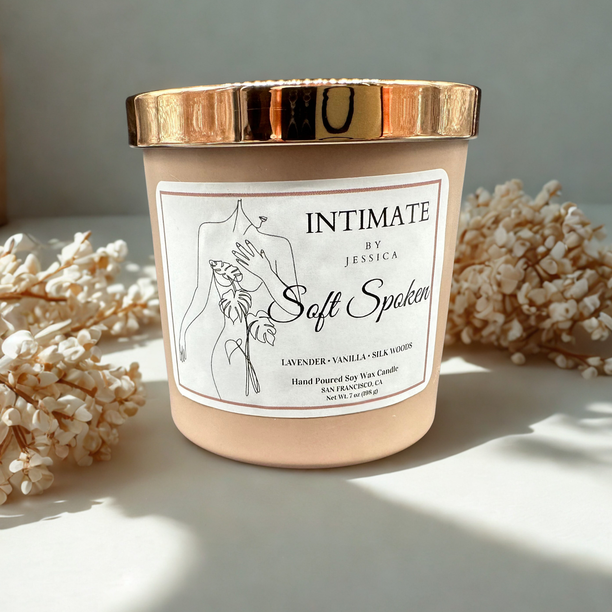 A beige candle container with a metallic gold lid, labeled "Intimate" by Jessica, with scent notes of lavender, vanilla, and silk woods, surrounded by white dried flowers.