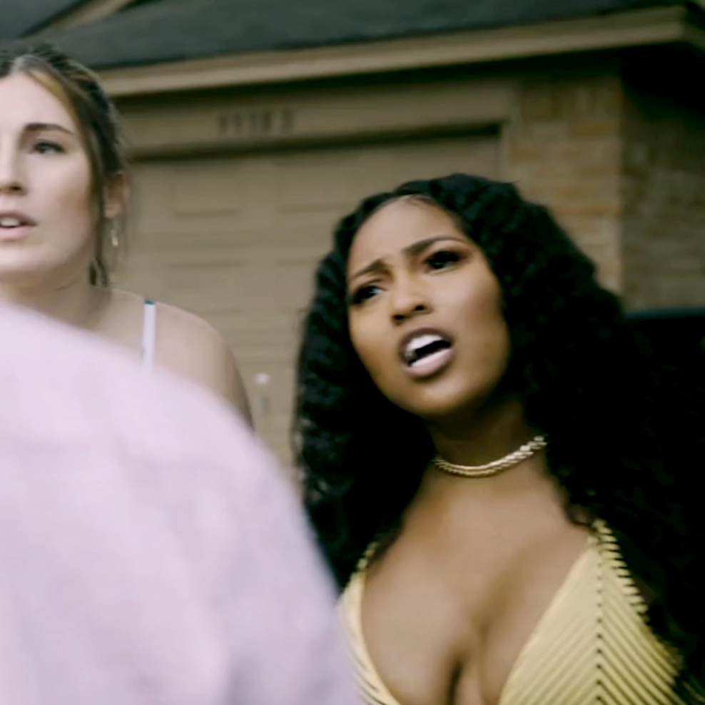 Two women having a conversation outdoors in front of a brick house; one woman with curly hair and a yellow top appears upset or confused, while the other with light hair looks on.