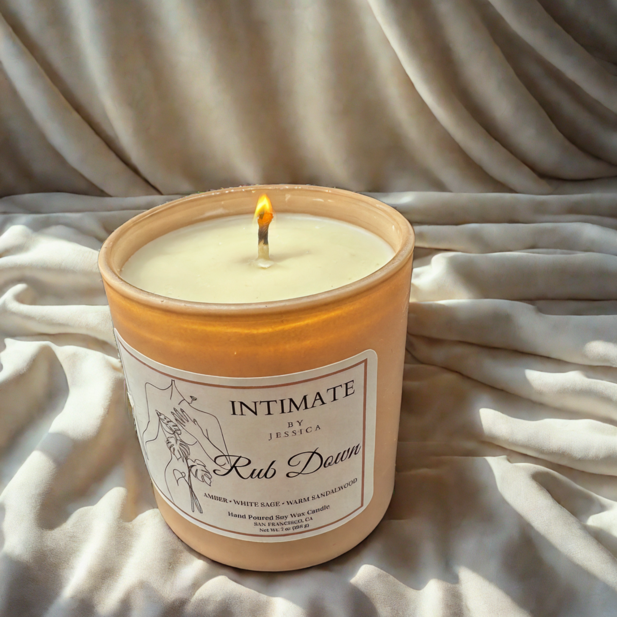 A lit scented candle in a ceramic holder with a label showing ingredients and scent details, placed on a fabric surface with a textured background.
