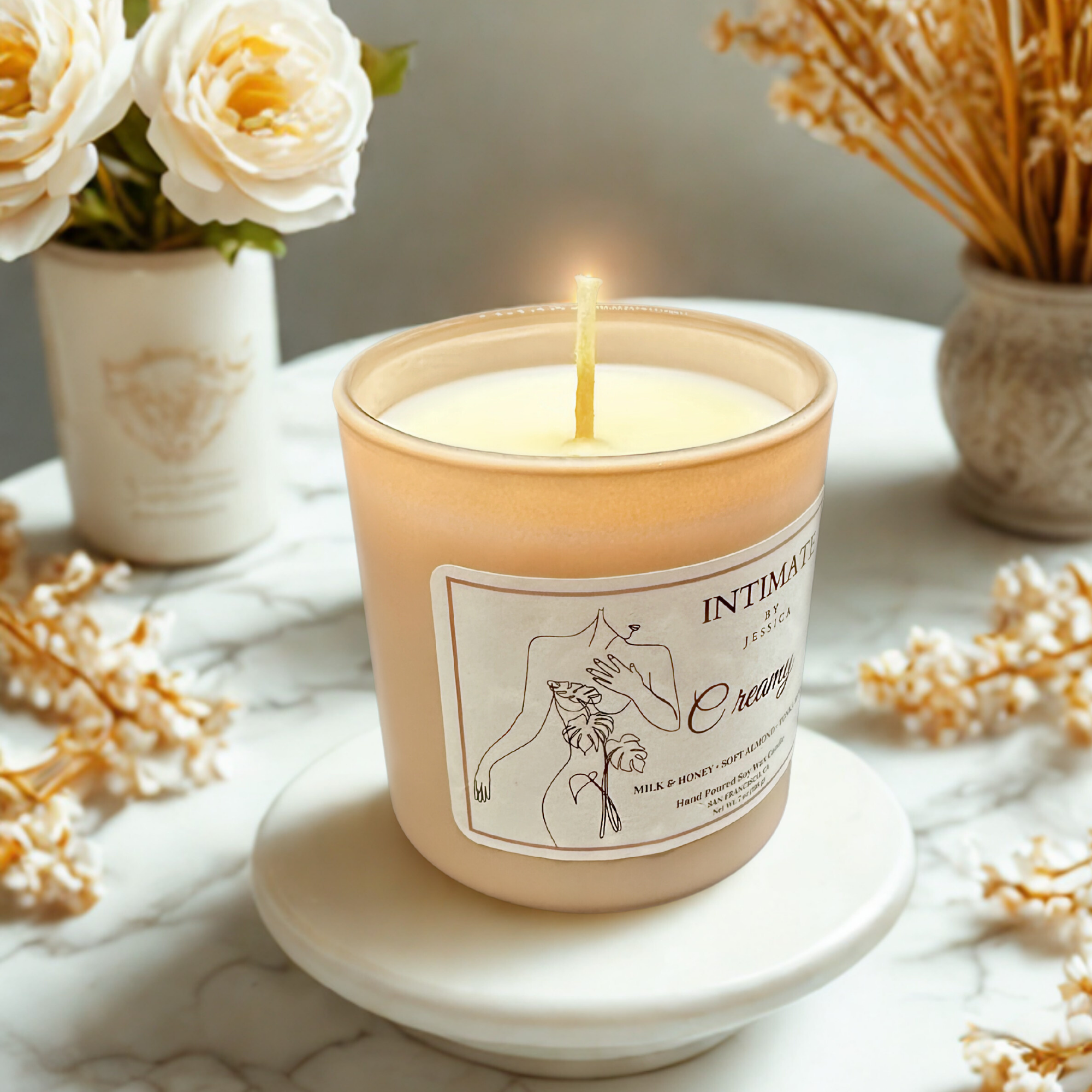 A peach-colored candle with a label featuring an illustration of a hand holding a plant, surrounded by white flowers and dried wheat stalks on a white surface.