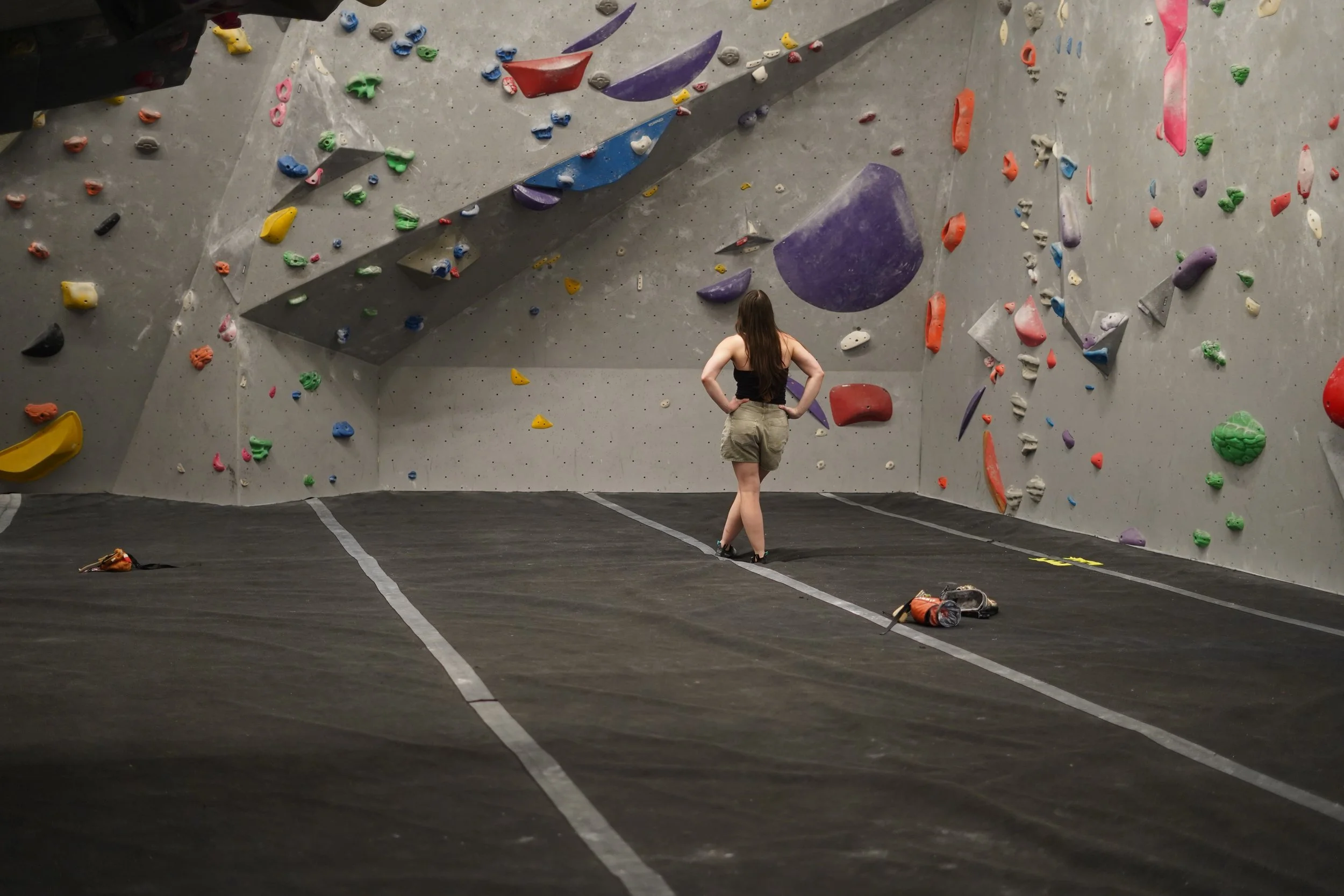 A woman standing in front of a large indoor climbing wall with colorful holds.