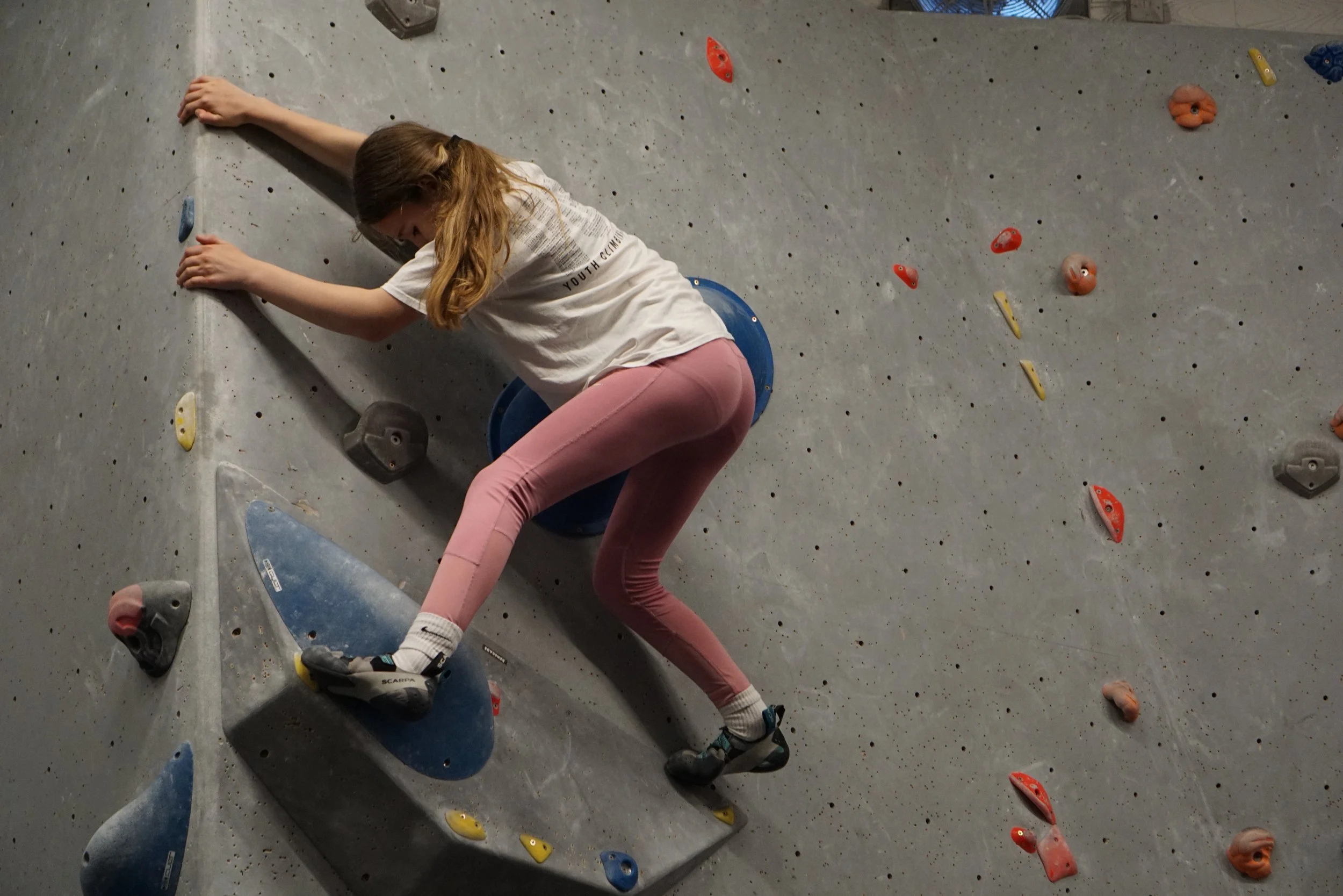 A young girl climbing an indoor bouldering wall with various colorful holds, wearing a white t-shirt, pink pants, and climbing shoes.
