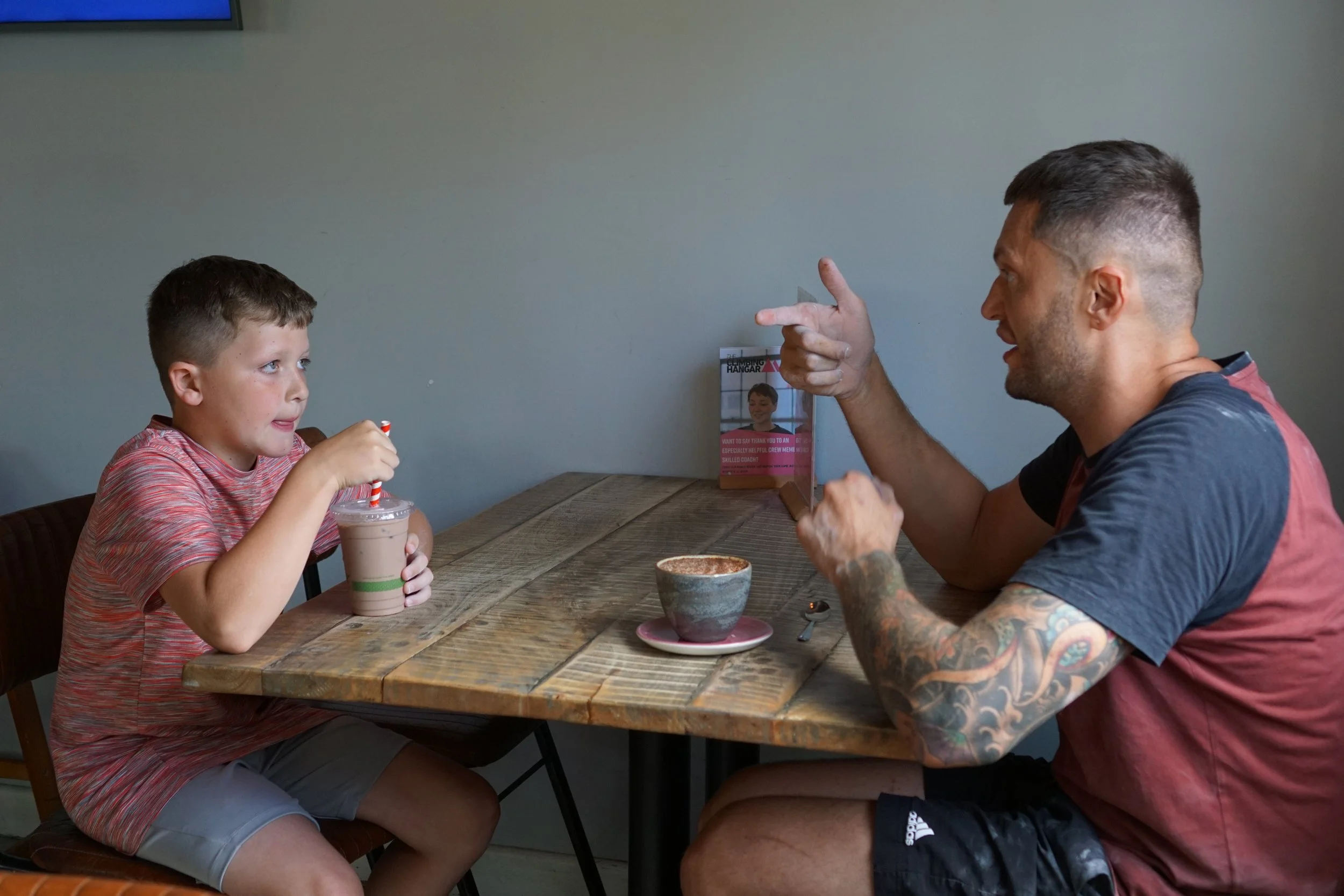 A young boy and an adult man sitting at a wooden table having a conversation. The boy is holding a drink with a red and white striped straw, and the man is gesturing with his hand. There are coffees on the table.