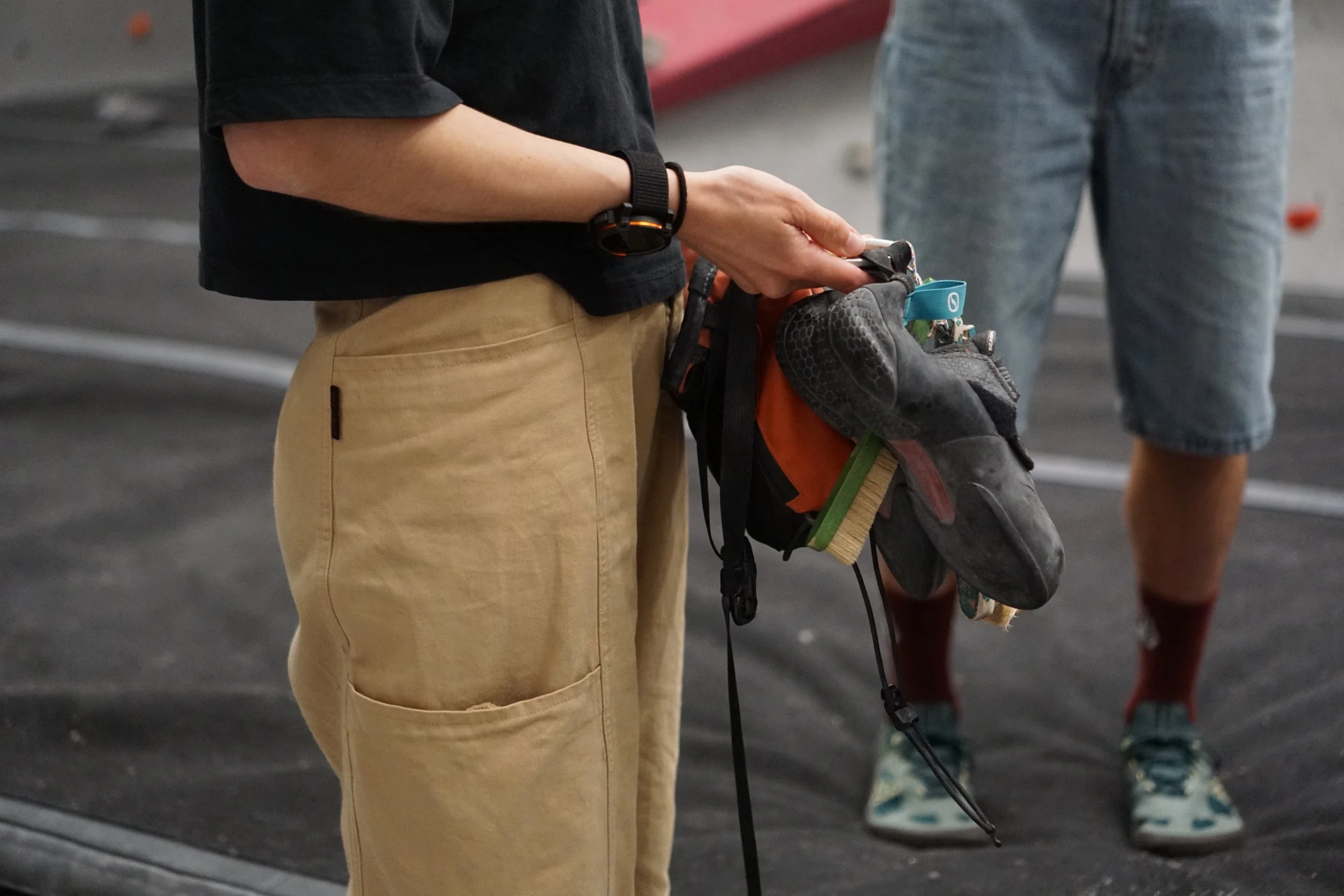 A person wearing a black T-shirt and khaki pants holding climbing shoes with a chalk bag, standing on the climbing mats.