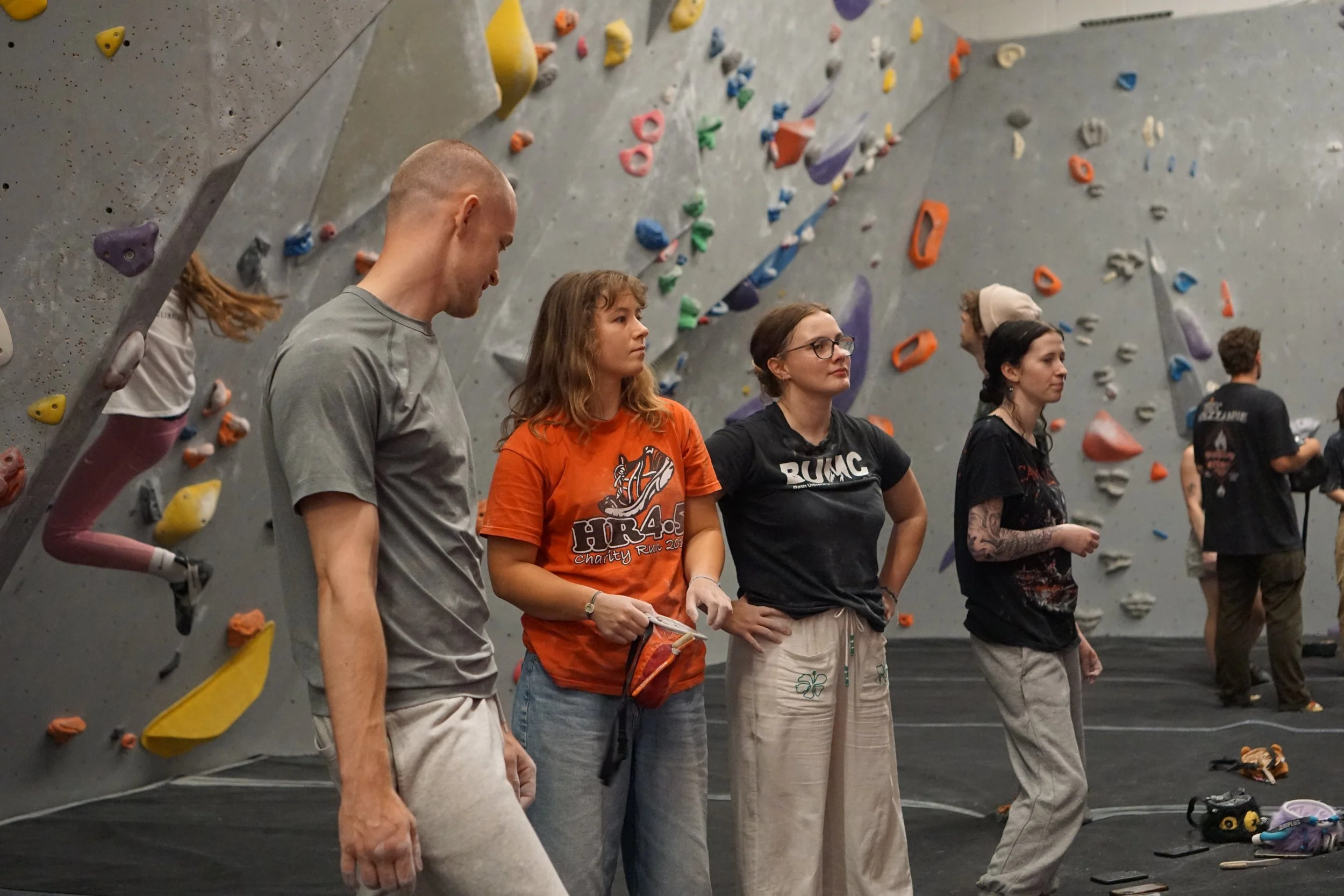Group of people inside an indoor rock climbing gym, standing on the ground with climbing holds on the wall behind them.