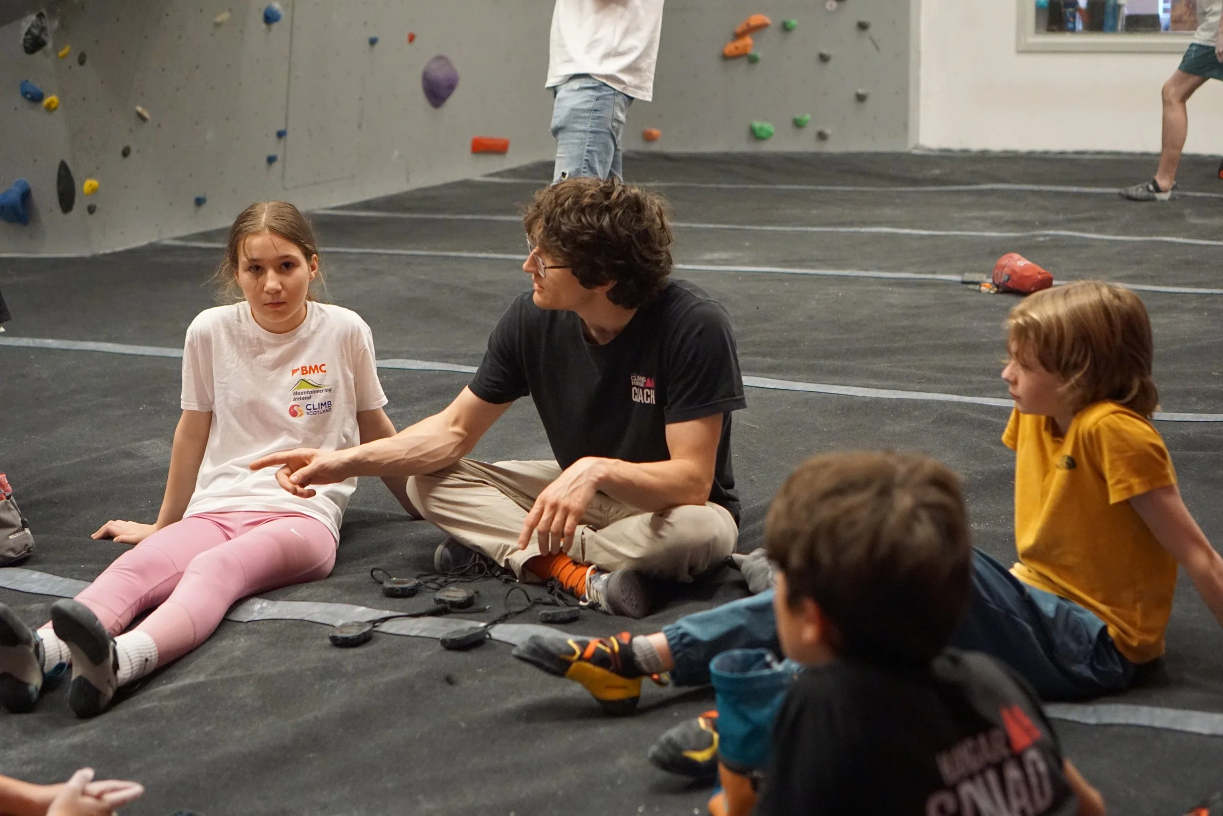 Children and an coach sitting on the ground in an indoor climbing gym, with rock climbing holds on the wall in the background.
