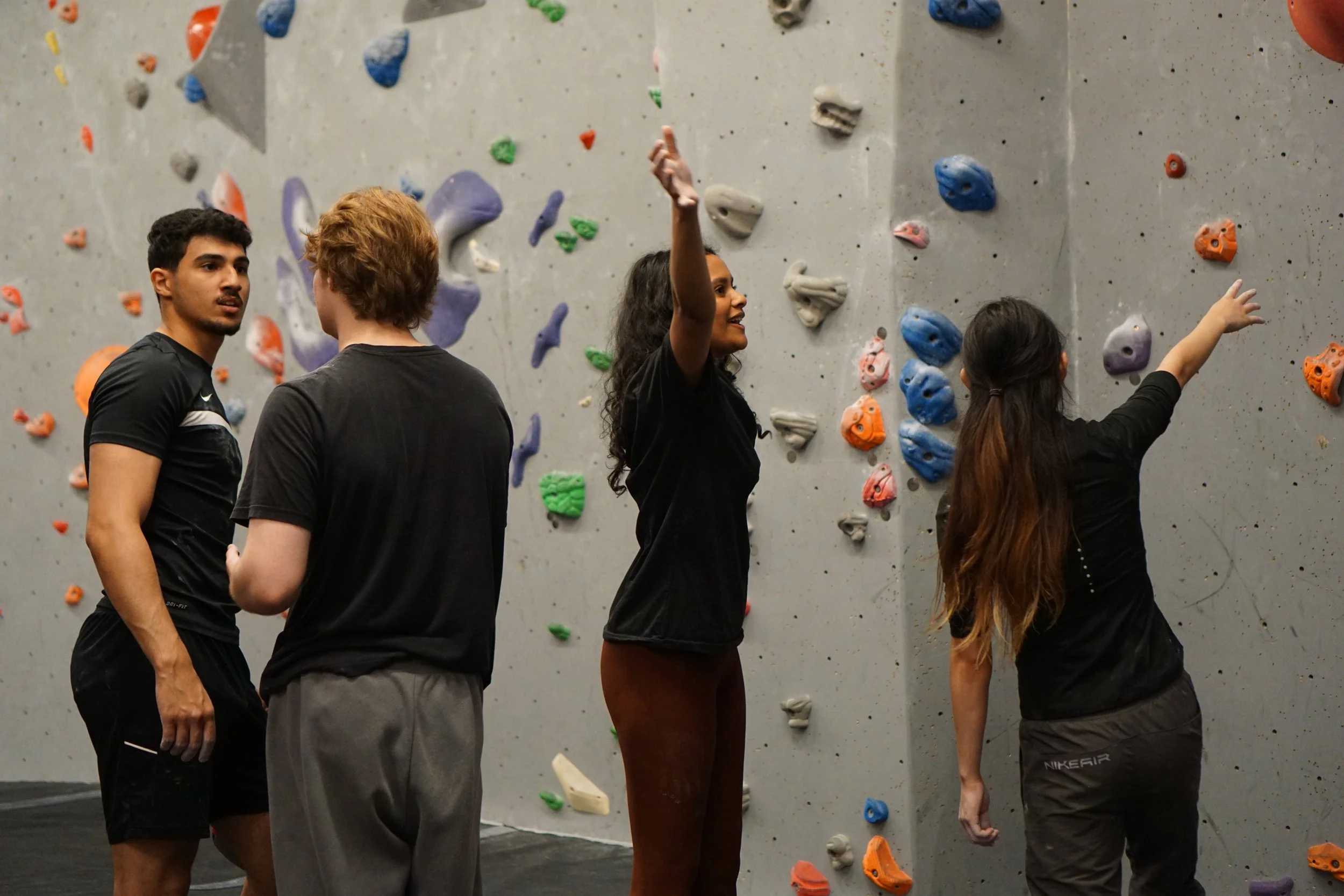 Four new climbers standing in front of an indoor climbing wall, engaging in conversation or training. The wall has various colourful holds.