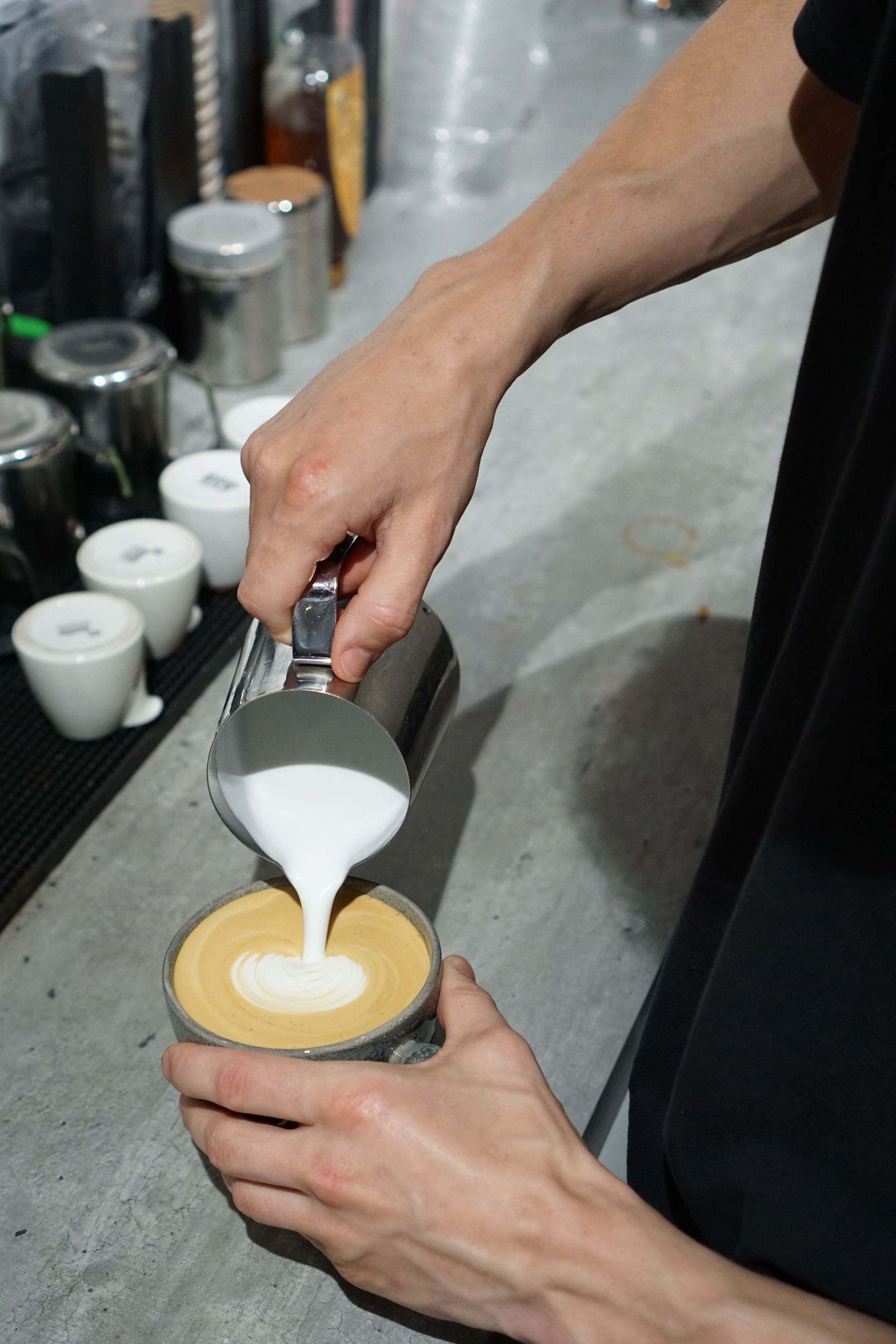 A person pouring milk into a cup of coffee to create latte art, with several coffee cups and containers on a counter in the background.