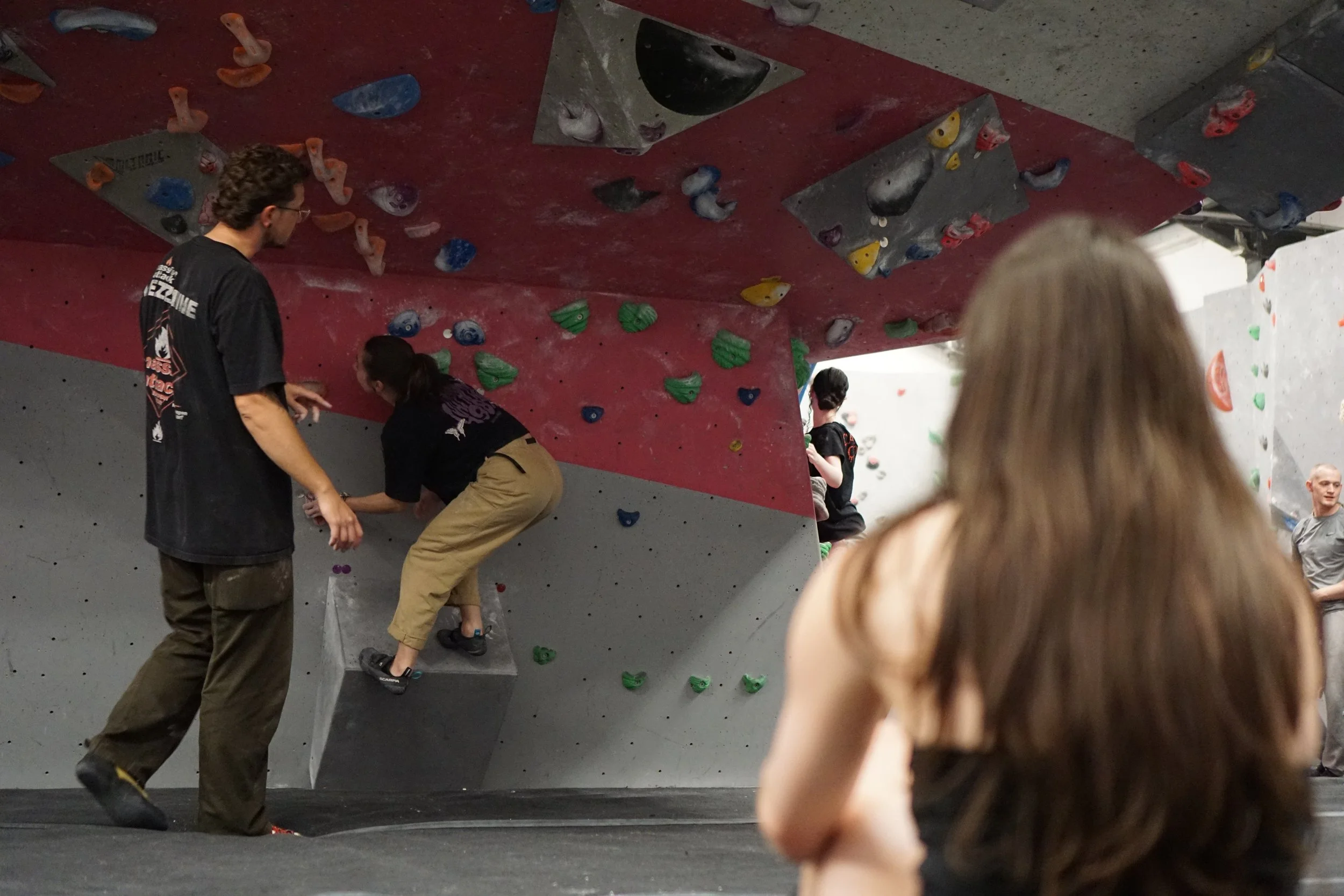 People inside an indoor rock climbing gym, preparing to climb, on a colourful climbing wall with various holds.