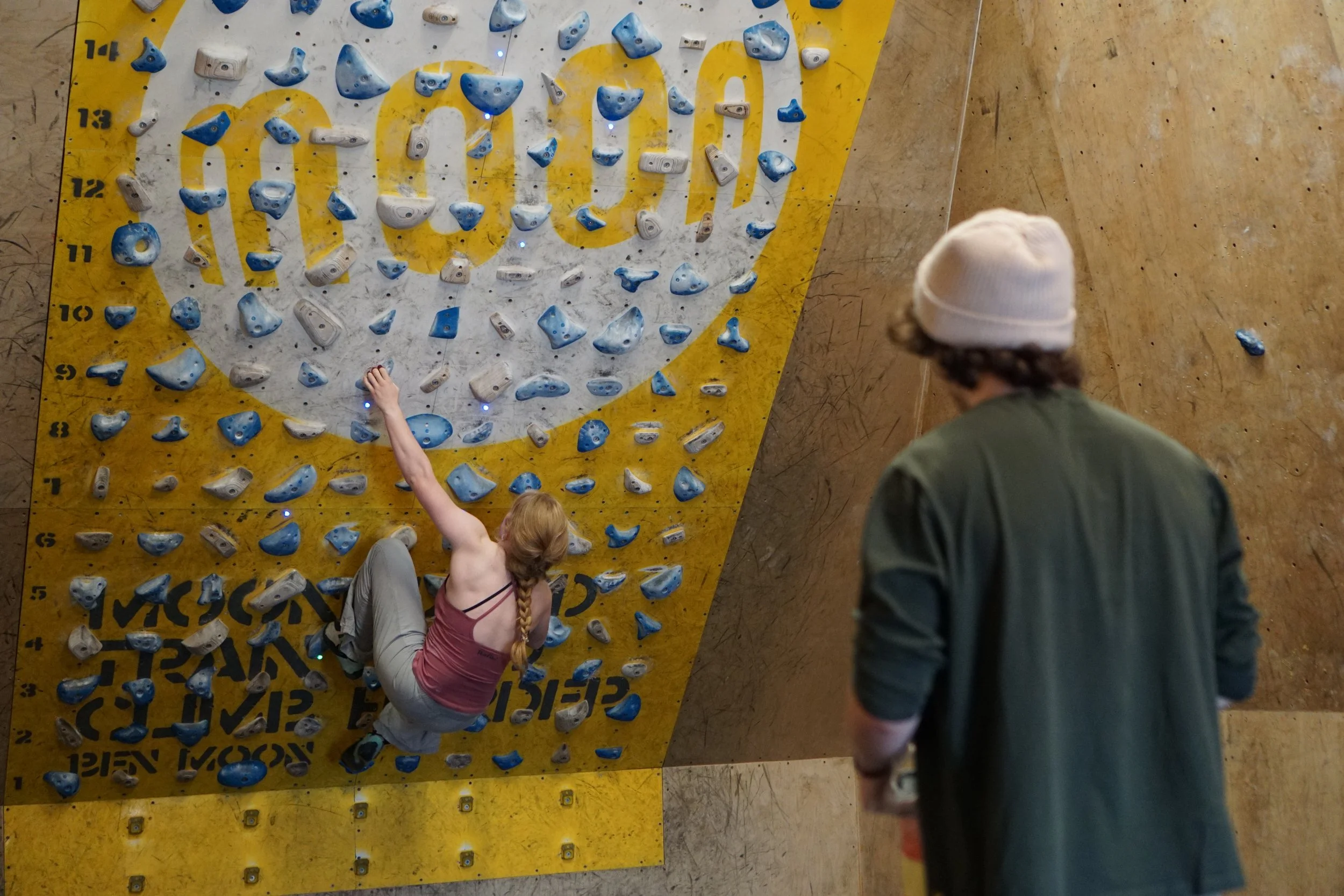 A woman with red hair in a braid is climbing an indoor rock climbing wall with a friend watching nearby. The wall is a Moonboard