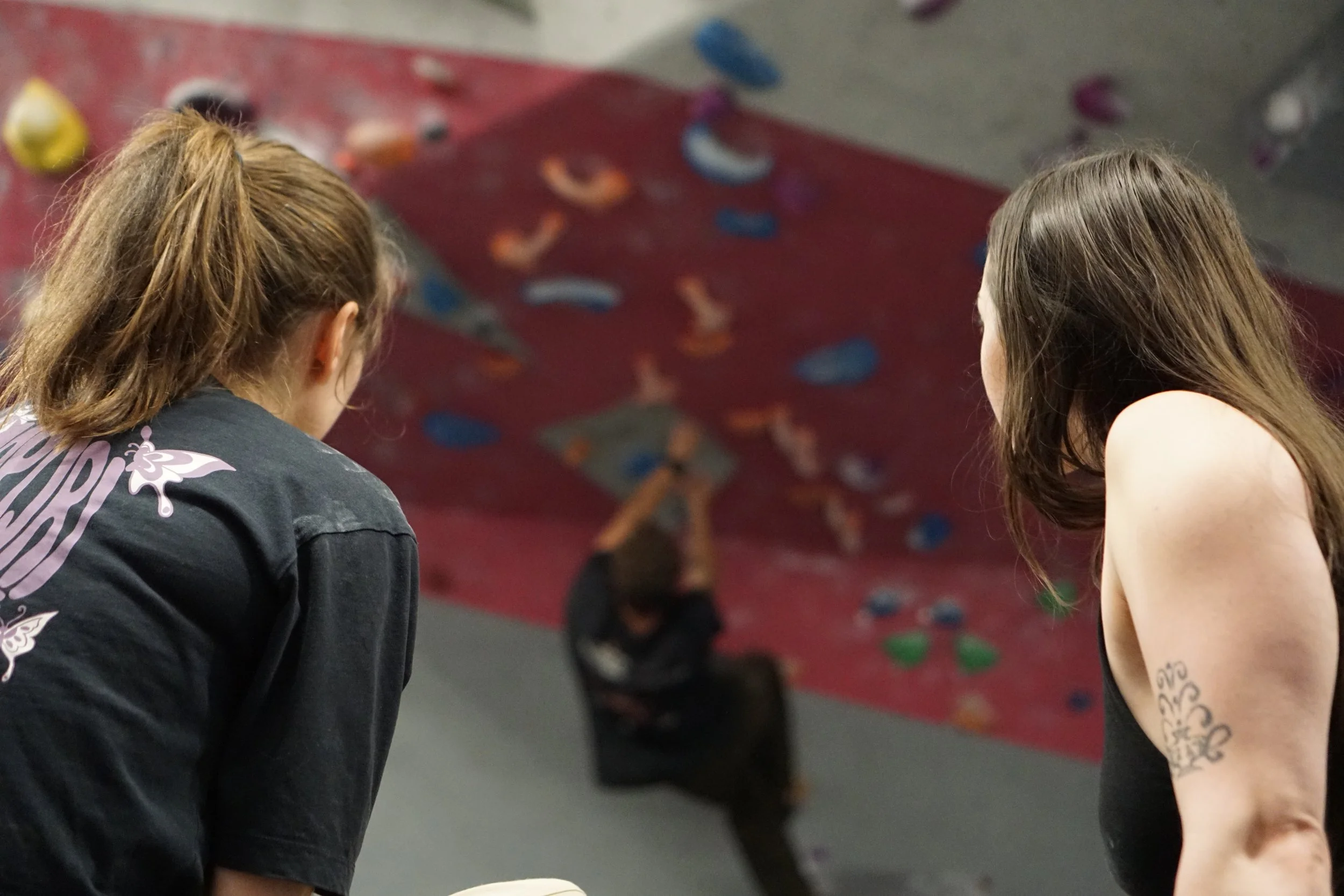 Two women with long hair, one with a ponytail, wearing casual clothes, are watching an indoor rock climbing wall with colorful holds.