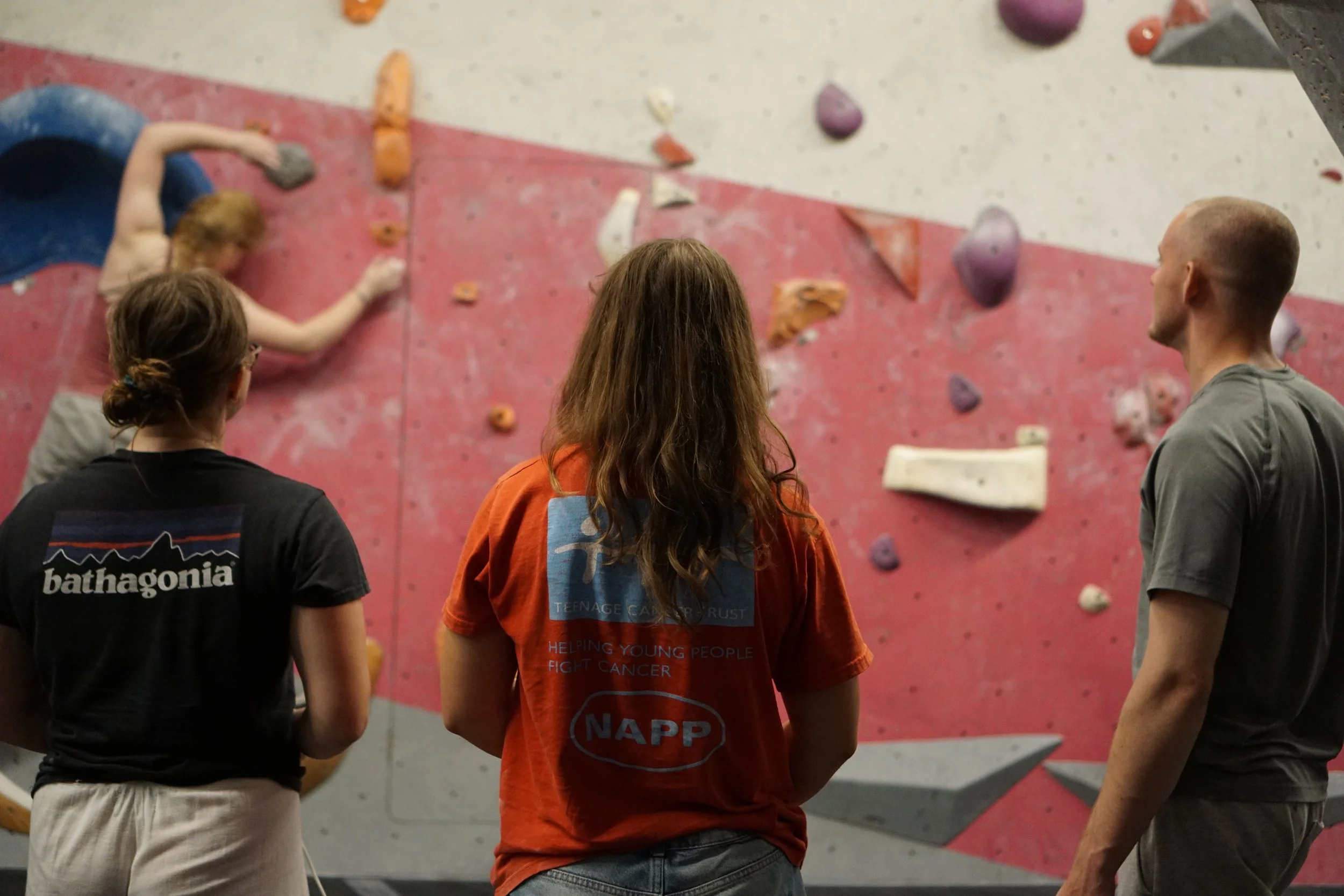 Four people stand in front of an indoor climbing wall, observing a climber. The climber, a young girl, is reaching for a hold on the wall. The others watch her, and the wall has colorful holds and grips.