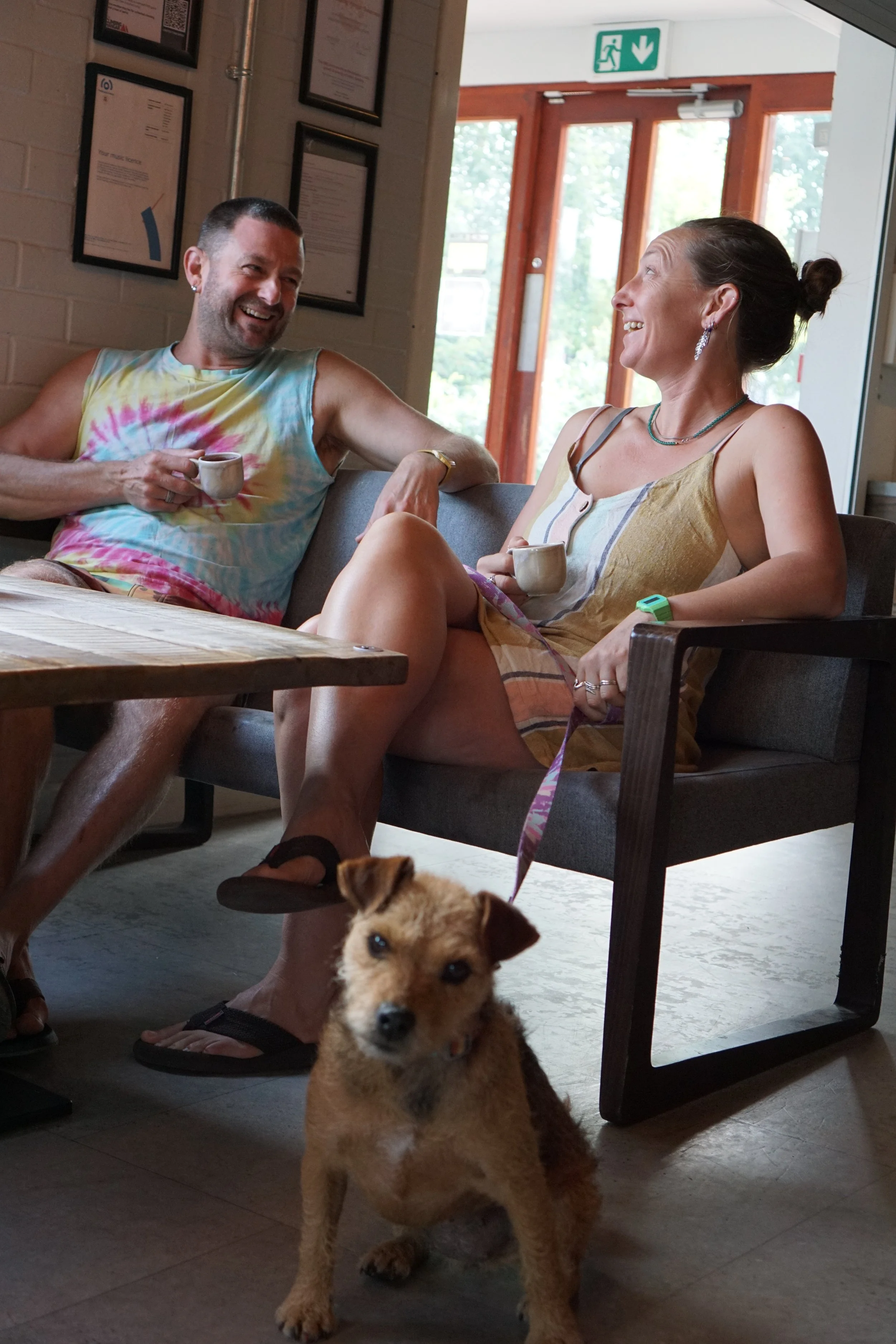 A man and woman sitting on a couch inside The Hive cafe, laughing and enjoying coffee, with a small brown dog (Zavvi) standing in the foreground.