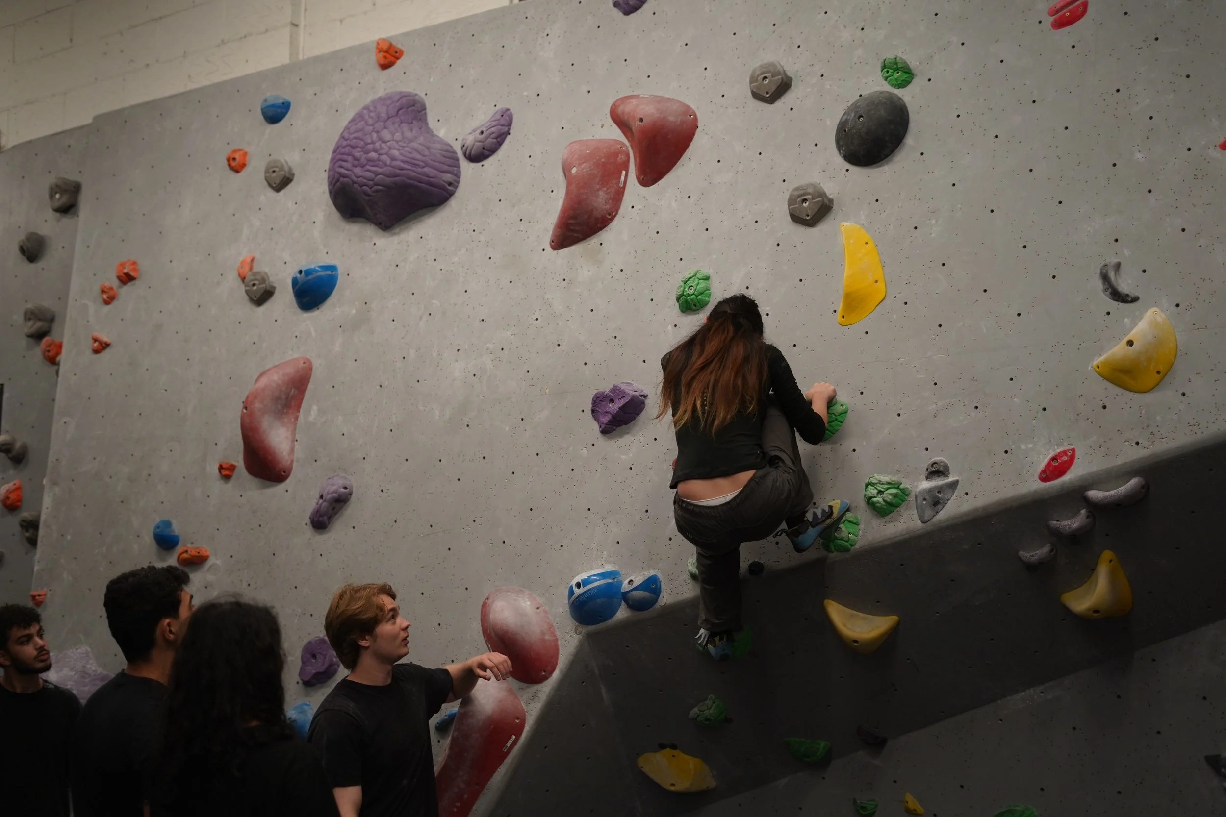 A group of people at an indoor rock climbing gym. One person is climbing the wall while others watch. The climbing wall is covered with colorful holds of varying shapes and sizes.