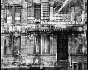 Black and white photo of a building's façade with barred windows, a balcony, and a person taking a photo reflected on a car window.