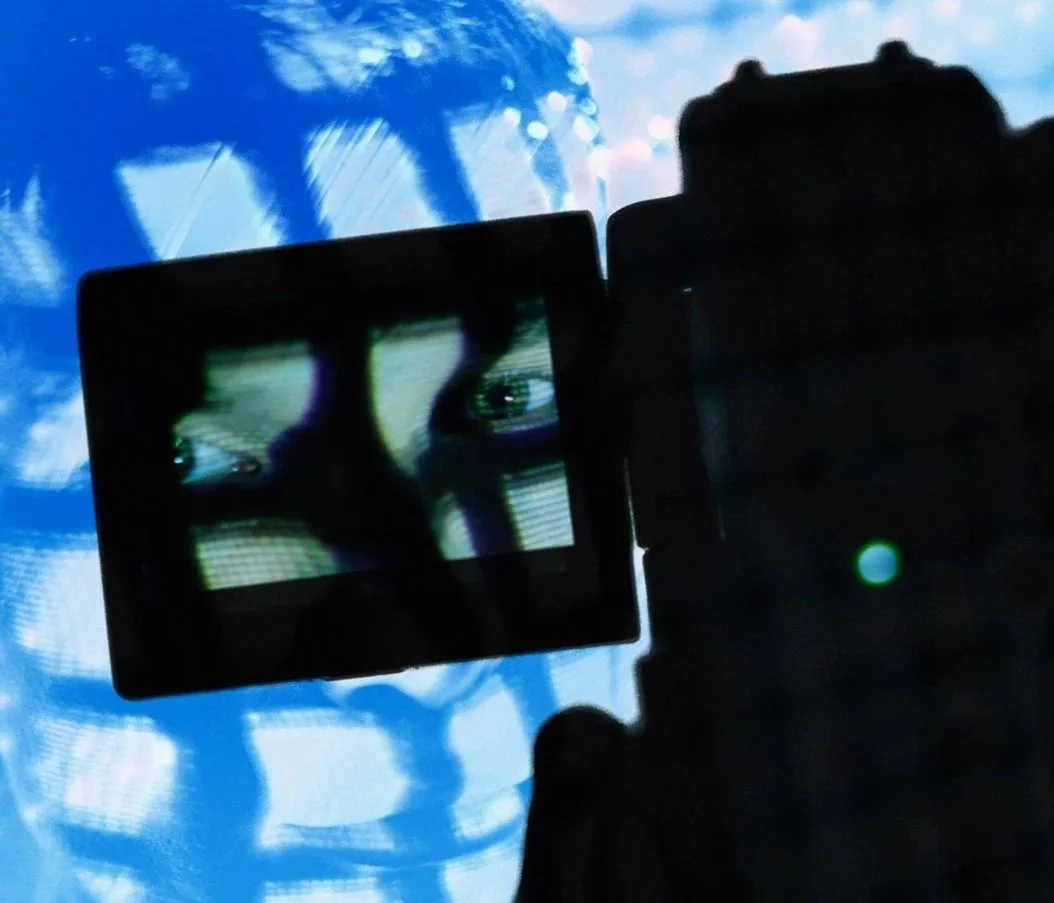 A person is taking a photo of their reflection on a camera screen, which shows their face, against a background of blue sky and clouds.