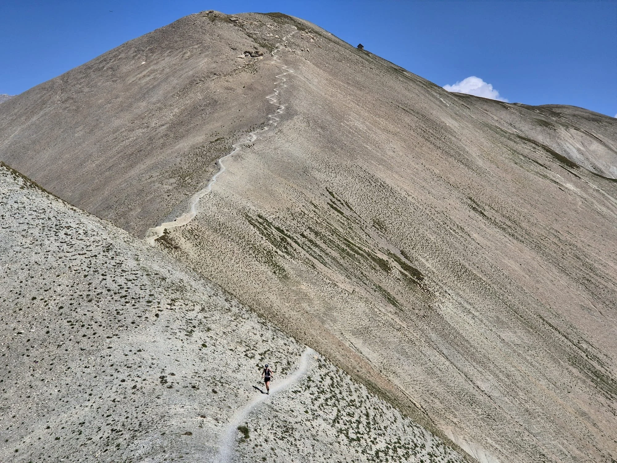 A hiker walking on a narrow mountain trail near the summit of a steep, rocky mountain under a clear blue sky.