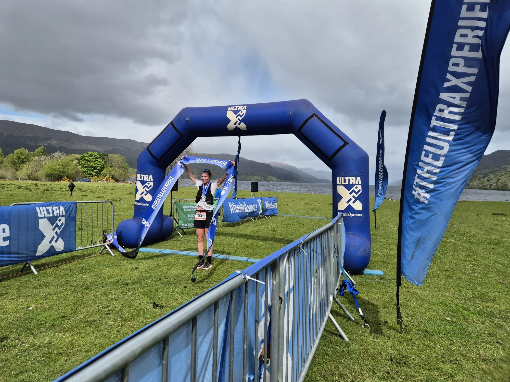 A woman standing next to a blue inflatable finish line arch with 'ULTRA X' branding, holding a ribbon, at an outdoor race event near a lake with mountainous scenery in the background.