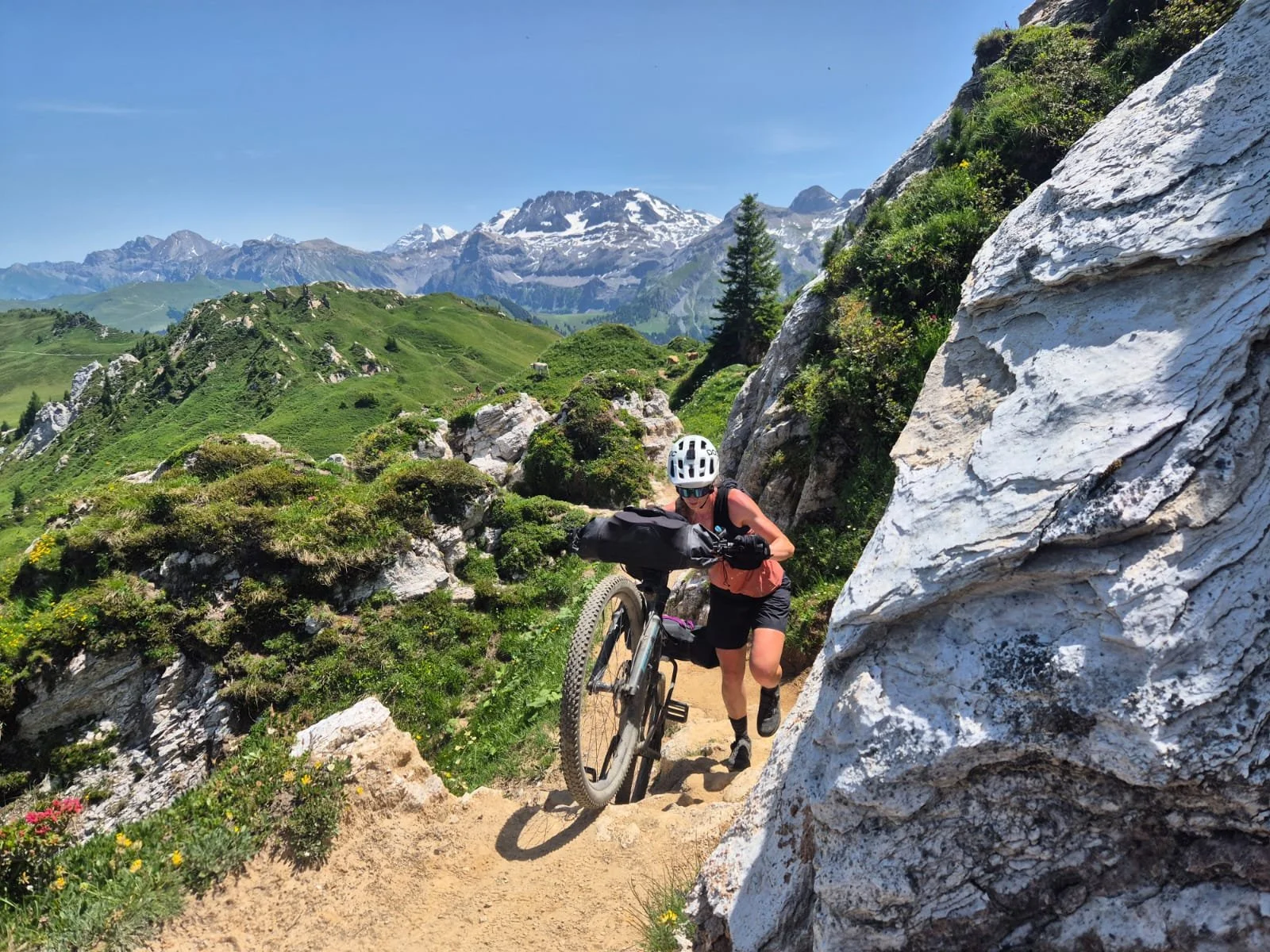 A person mountain biking on a narrow trail surrounded by green hills, rocky terrain, and snow-capped mountains under a clear blue sky.