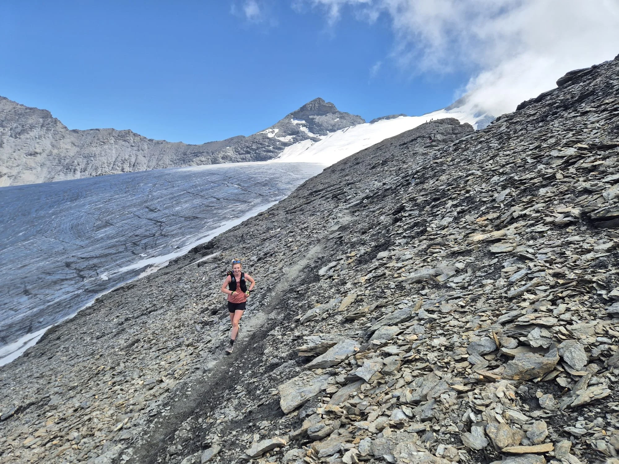 A woman in hiking gear running on a rocky mountain slope with snow and ice, with mountainous terrain and a partly cloudy blue sky in the background.