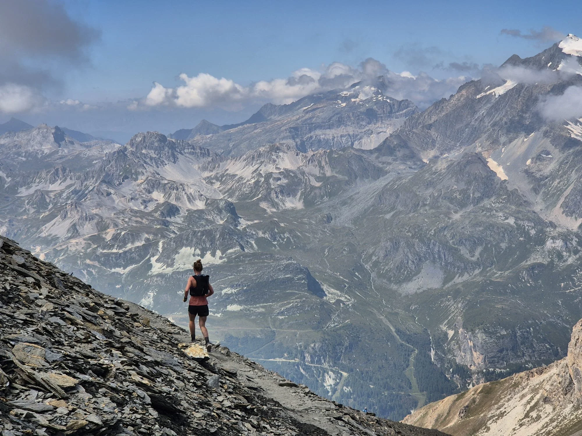 A person hiking on a rocky trail in a mountain landscape with snow-capped peaks and clouds in the background.