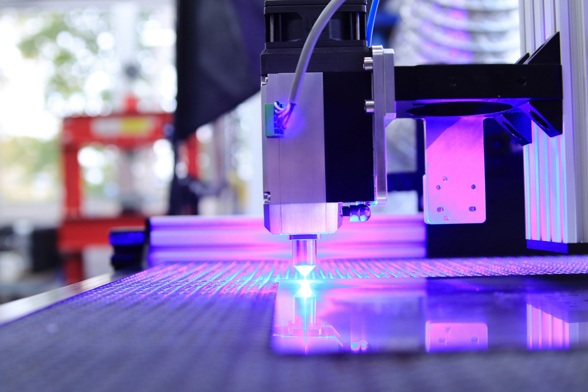 Close-up of a 3D printer nozzle extruding purple filament onto a build platform, with a blurred background of a workshop or laboratory.