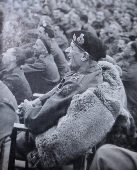 A black and white photo of military personnel, likely during a historical event, with a man in the foreground wearing a uniform, beret, and a thick fur coat, sitting among a group of soldiers or officers who are seated and appear to be listening or watching.