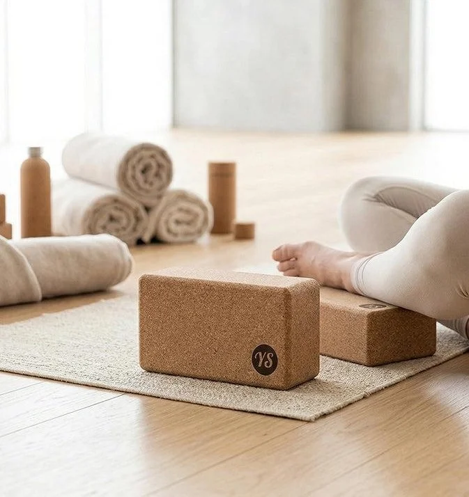 Person performing yoga or stretching on a cork yoga block in a peaceful indoor setting with rolled towels and yoga props in the background.