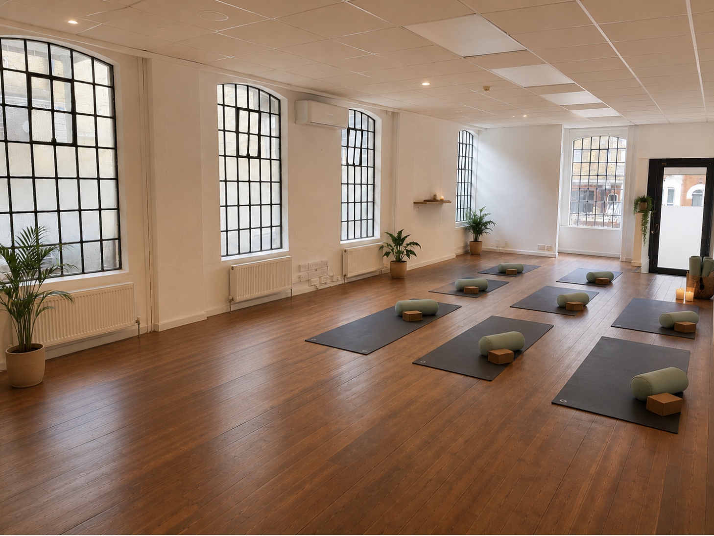 A spacious, well-lit yoga studio with wooden floors, large grid windows, and black yoga mats arranged in a circle. Each mat has a green yoga bolster and a wooden block. The room features potted plants, candles, and minimal decor, creating a calm and inviting atmosphere for yoga or meditation classes.